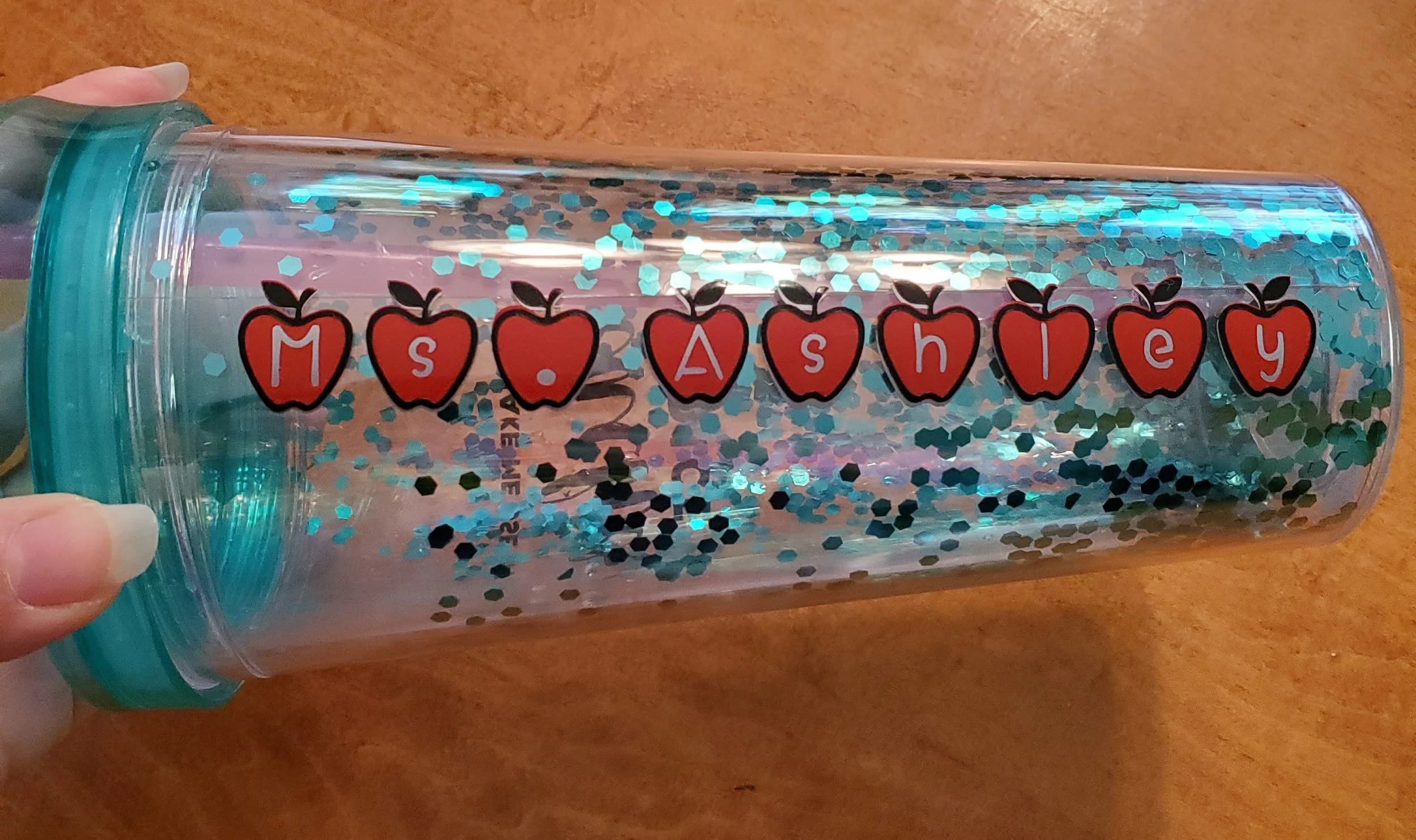 A clear plastic tumbler with blue glitter inside and a sticker of red apples with black leaves, spelling out 'Ms. Ashley' in white letters.