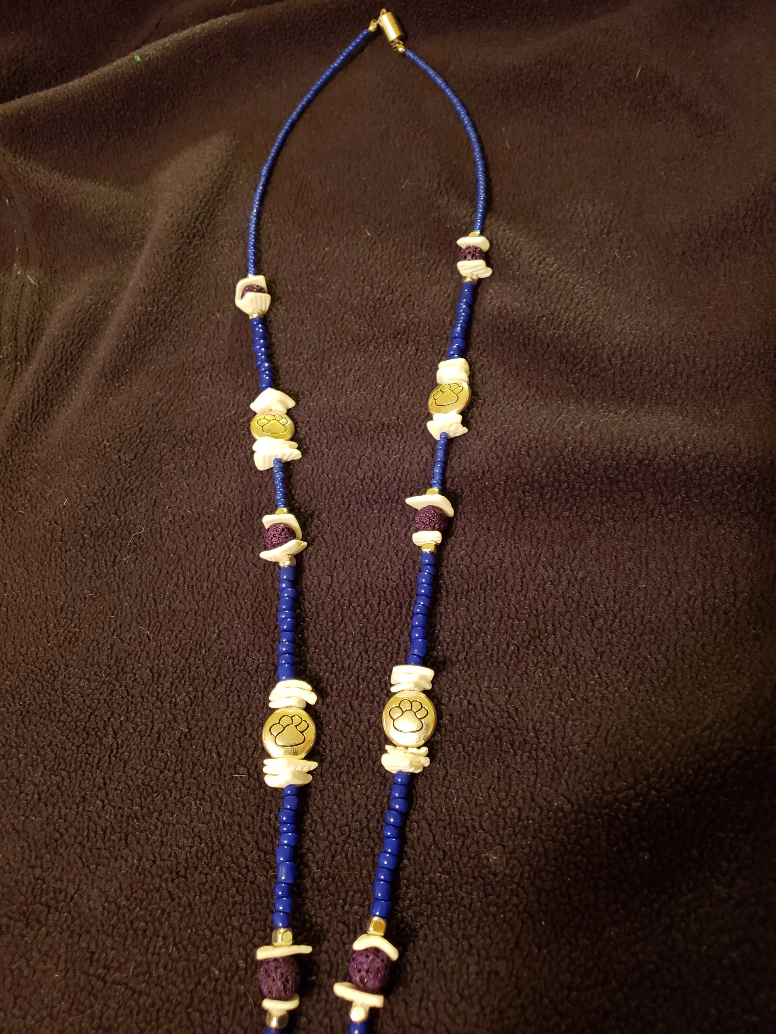 Long beaded necklace with navy blue, white, gold, and purple beads on a brown fabric background.