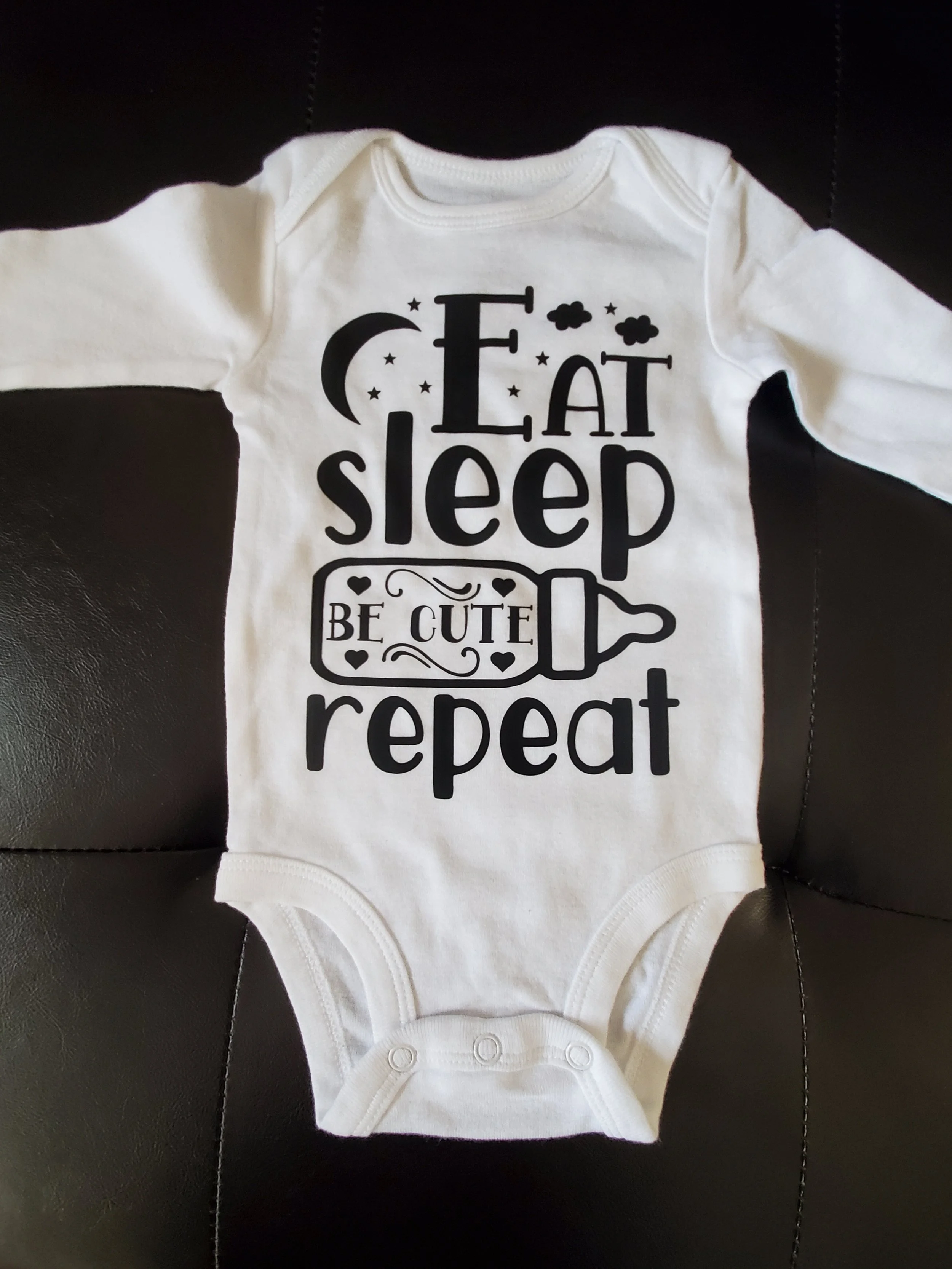 White baby onesie with black print text that says 'Eat sleep be cute repeat,' decorated with a moon, stars, clouds, and a baby bottle