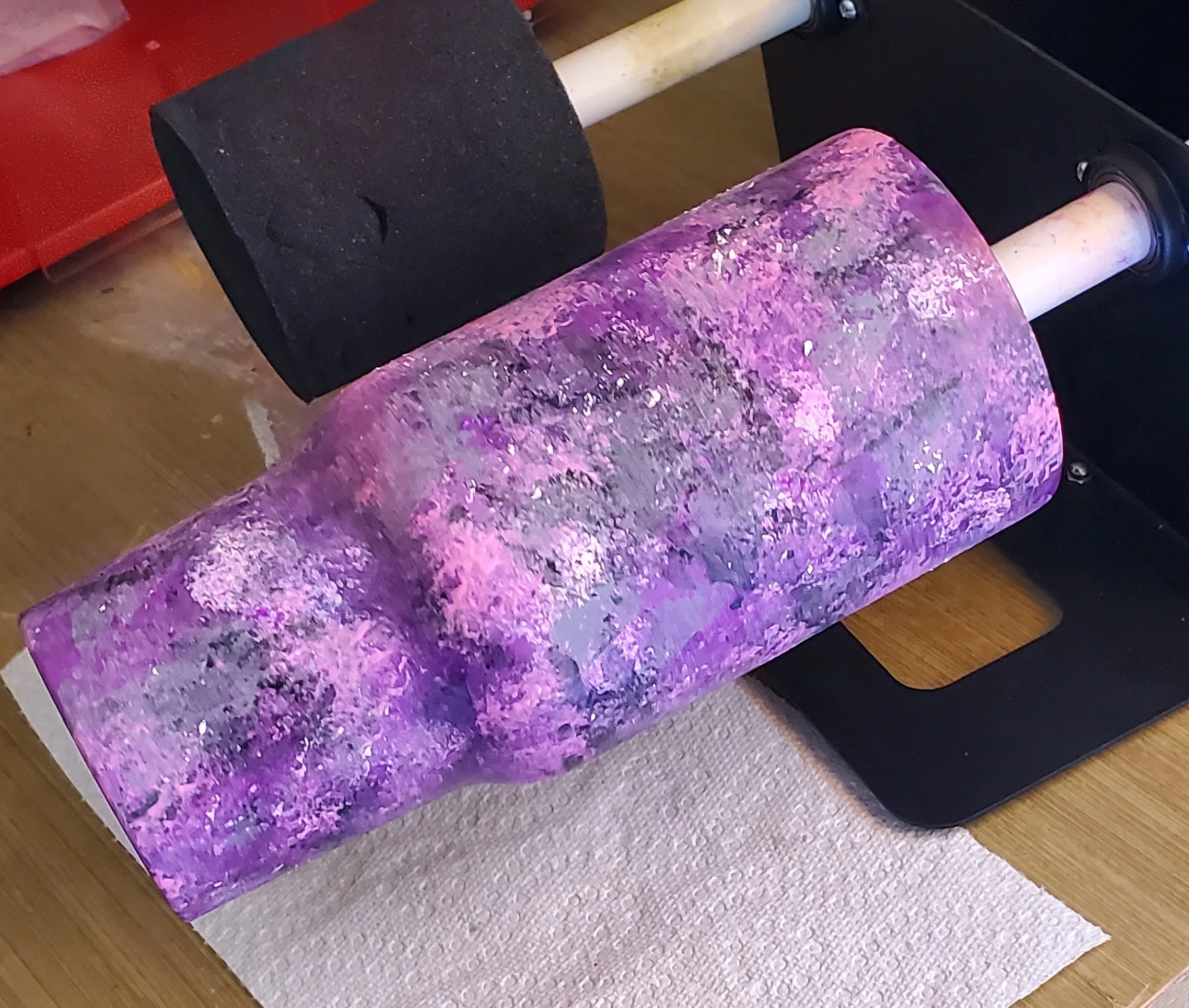 A cylindrical object painted to resemble a galaxy with pink, purple, and white splatters, mounted on a black stand on a wooden table.