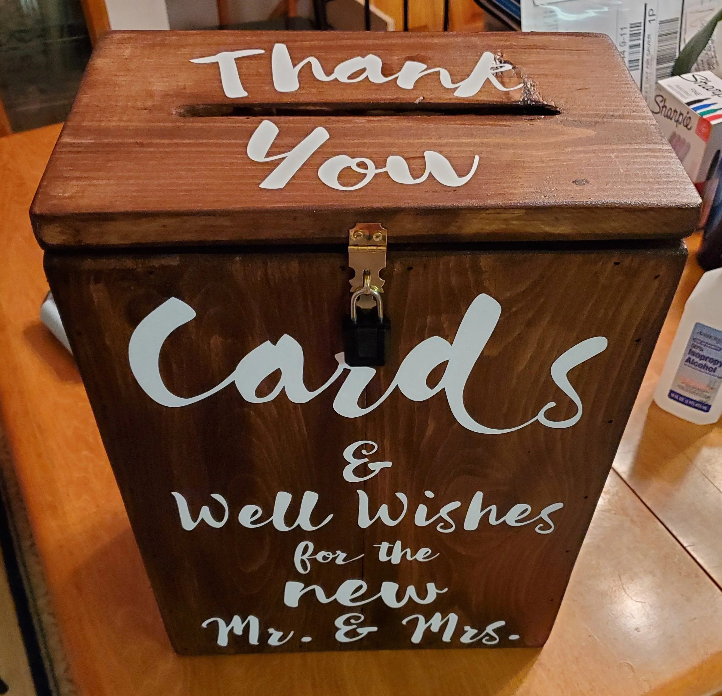 A wooden donation box with white lettering that says, "Thank You," "Cards," "& Well Wishes for the new Mr. & Mrs." and a lock on the front.