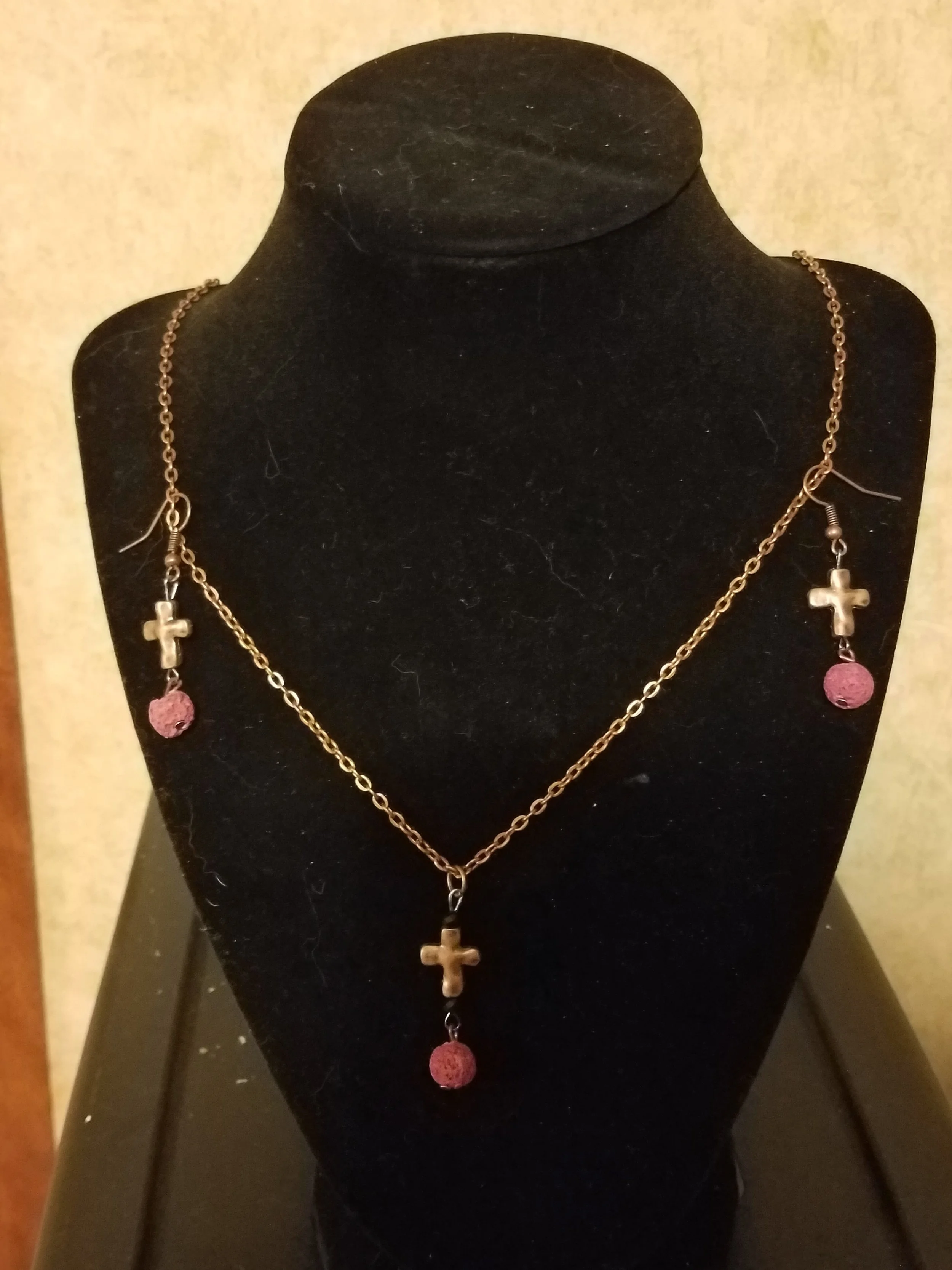 A gold necklace with three cross pendants and three pink beads displayed on a black velvet bust.