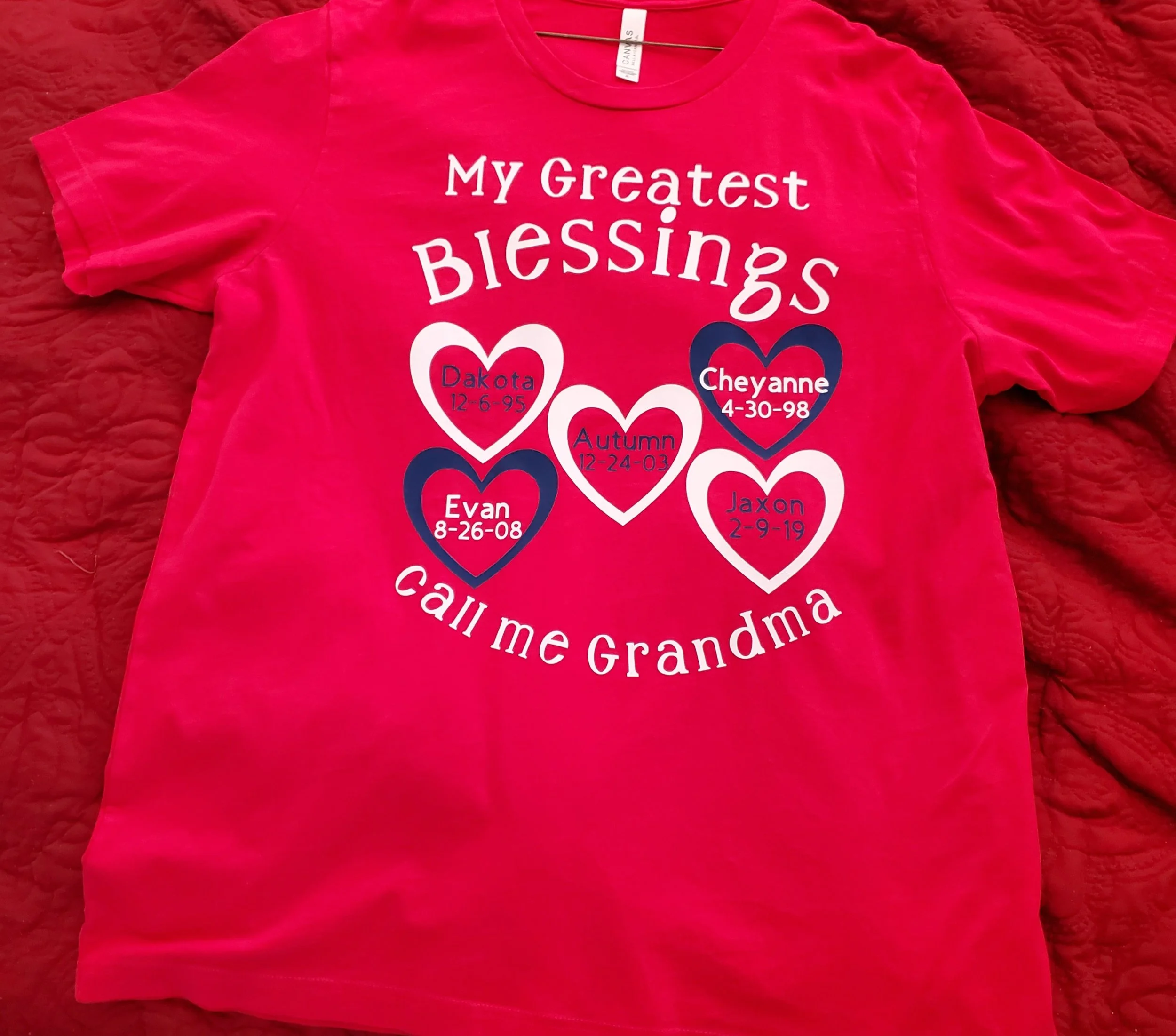 Pink T-shirt with white and dark blue text and hearts, reading "My Greatest Blessings" at the top and "call me Grandma" at the bottom. The shirt features five hearts with names and birth dates of grandchildren: Dakota (12-6-95), Autumn (12-24-03), Ev