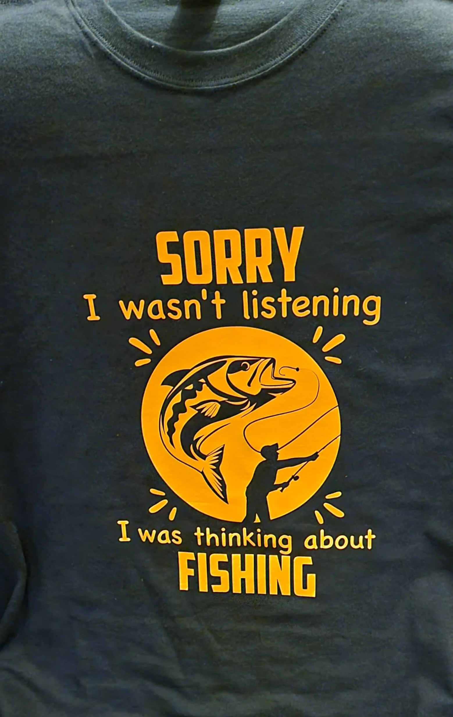 Black T-shirt with yellow graphic and text that says, 'Sorry I wasn't listening I was thinking about fishing,' featuring a person fishing and a large fish.