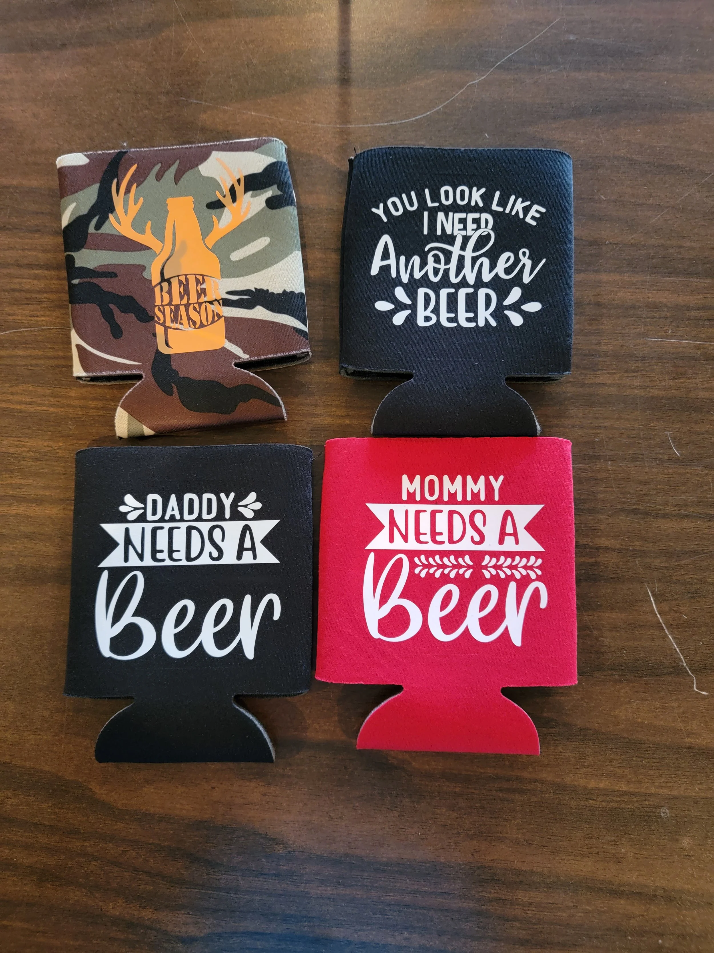 Four foam can holders with beer-themed quotes and designs on a wooden table.