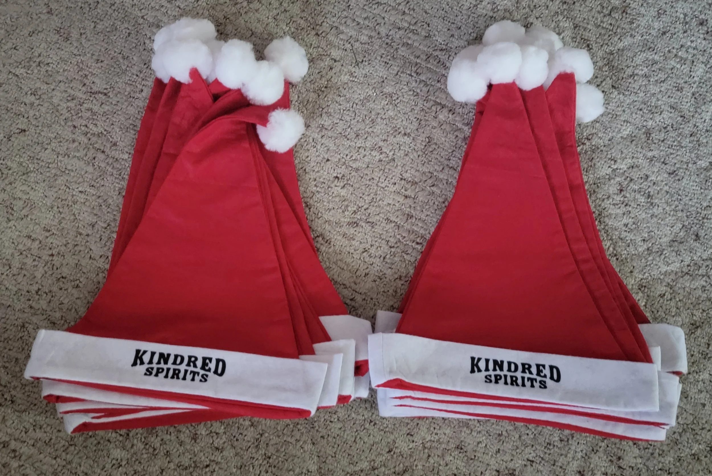 Two red Santa hats with white pom-poms and trim, labeled 'Kindred Spirits,' lying on a beige carpet.
