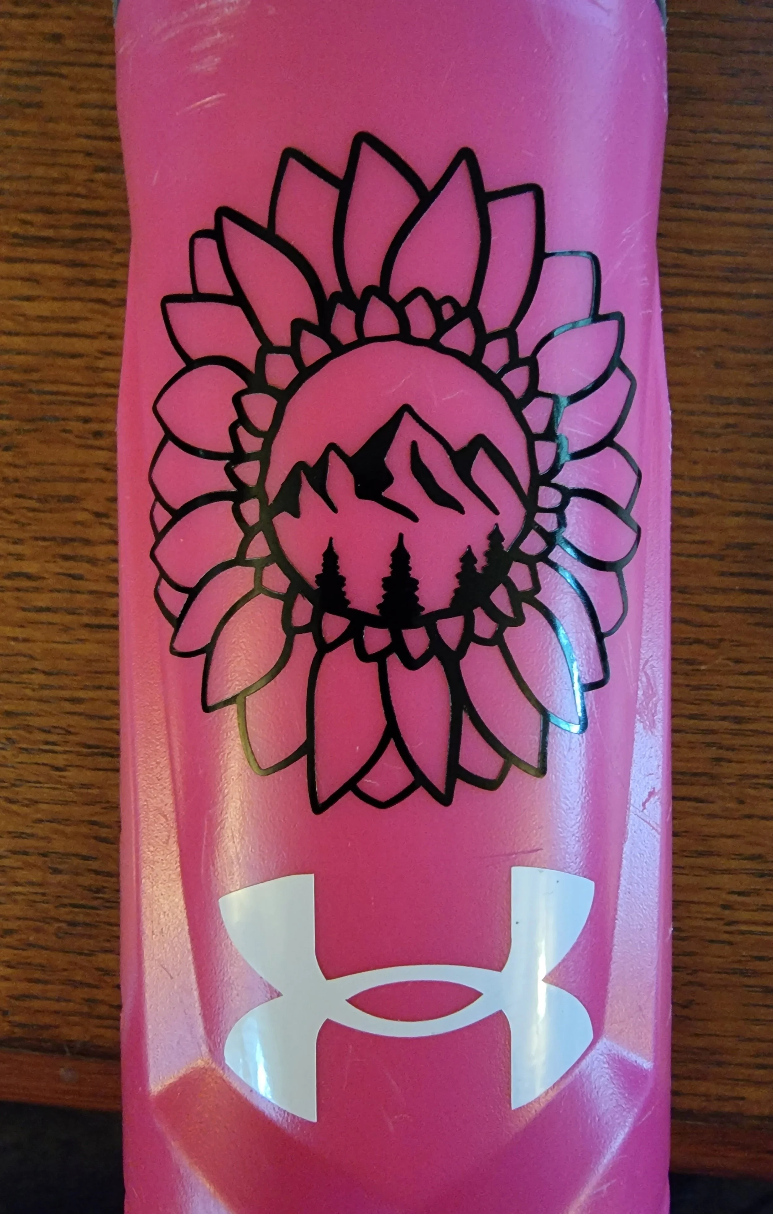 Pink water bottle with a black line drawing of a sunflower, mountains, evergreen trees, and a large white Under Armour logo.