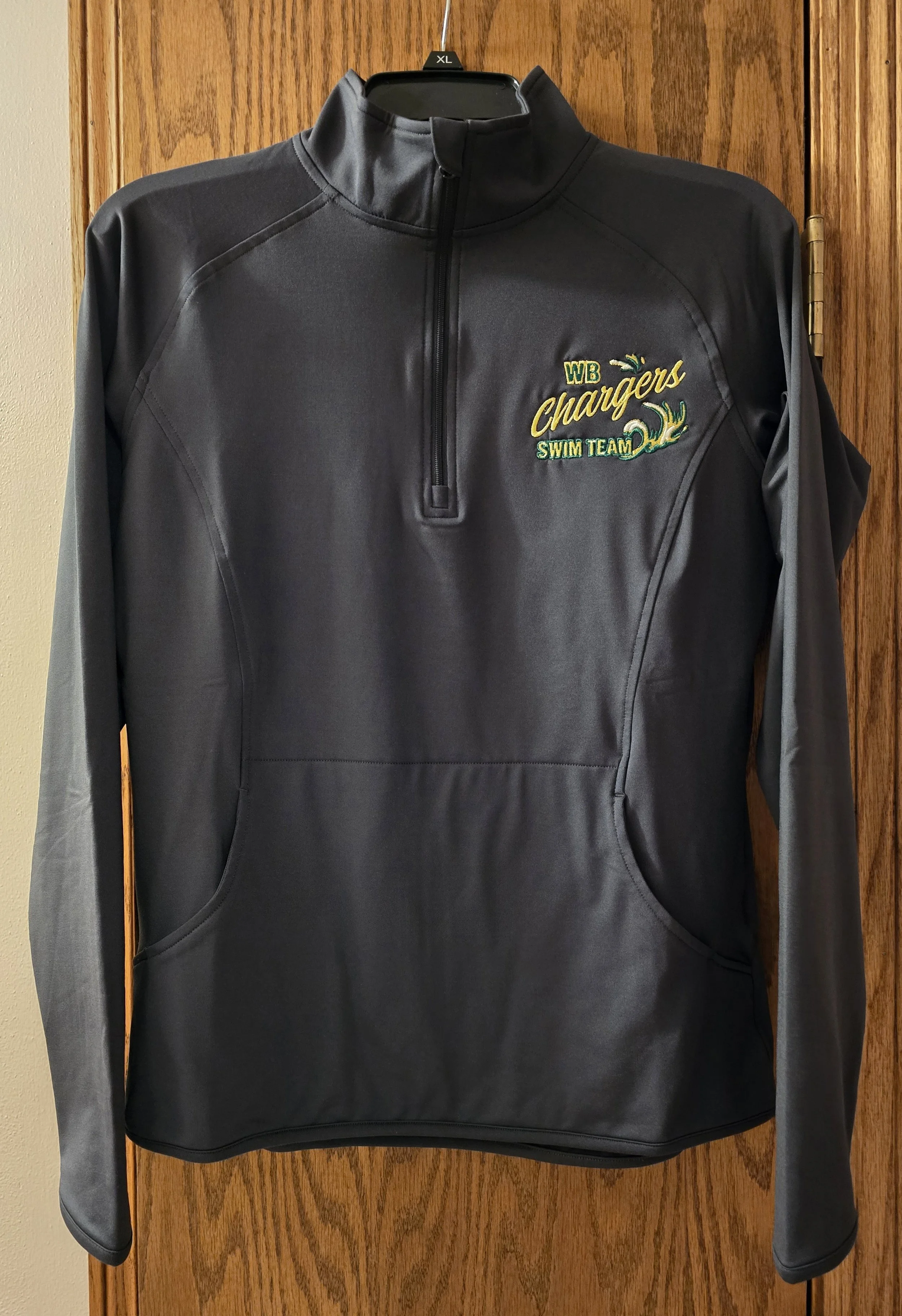 A black athletic quarter-zip pullover with a logo on the chest reading "WB Chargers Swim Team" and a stylized wave design, hanging on a black hanger against a wooden background.