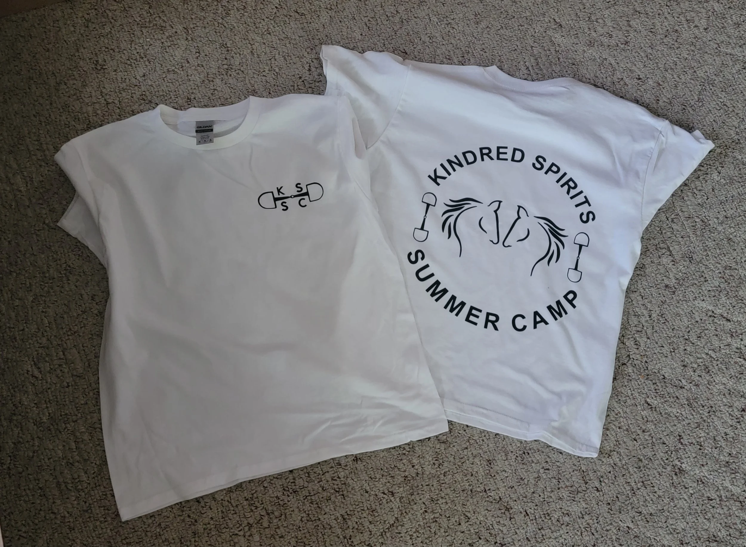 Two white T-shirts laid flat on a beige carpet. The front of one has a small skateboard graphic with the letters "K S C D". The back of the other has a circular design with a lion's face, the text "KINDRED SPIRITS SUMMER CAMP," and two small shovels.