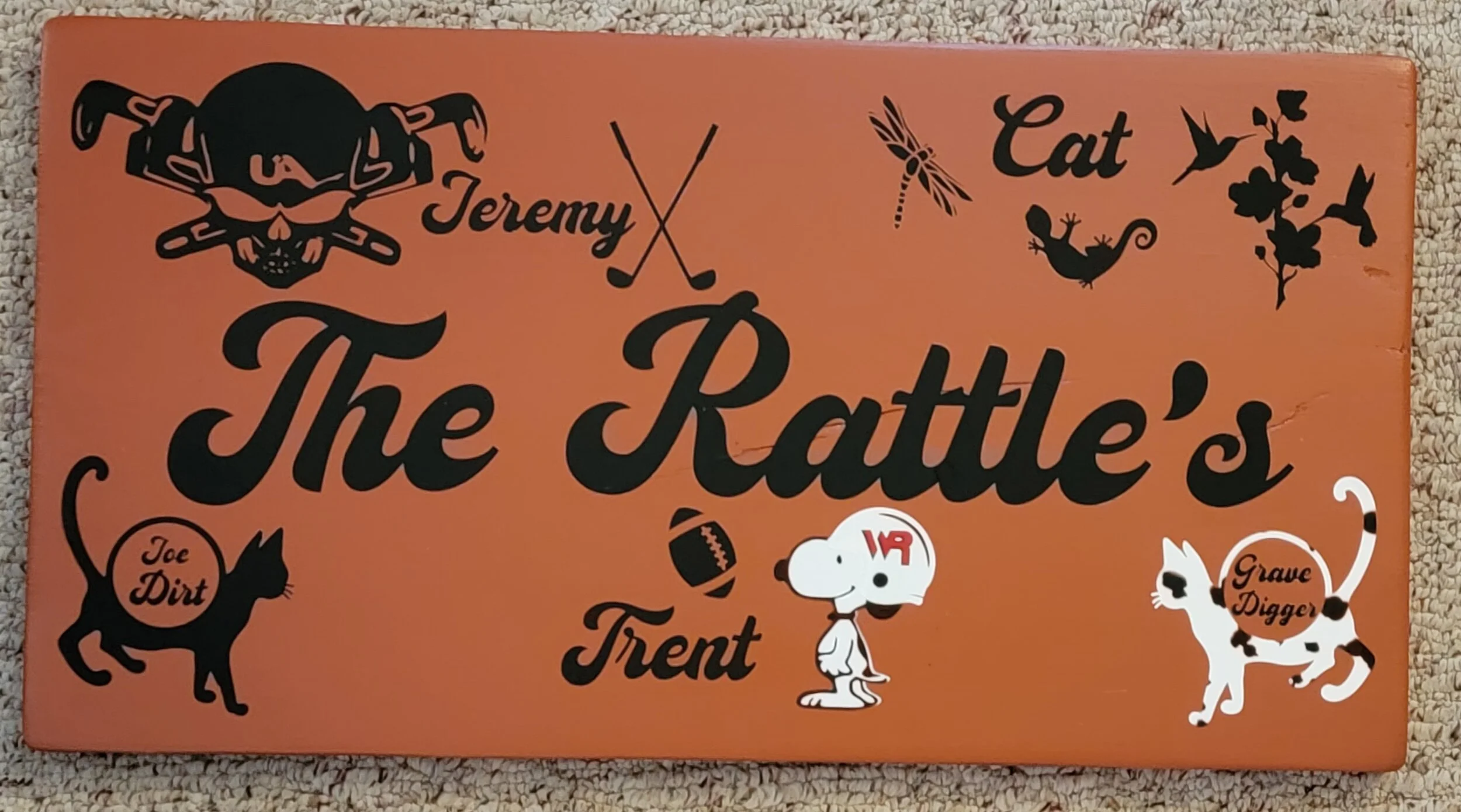Decorative sign with black illustrations and text on an orange background, reading "The Rattle's" with the names Jeremy, Trent, and Grave Digger, along with symbols of a skull, cat, dragonfly, lizard, football, and cartoon dogs.