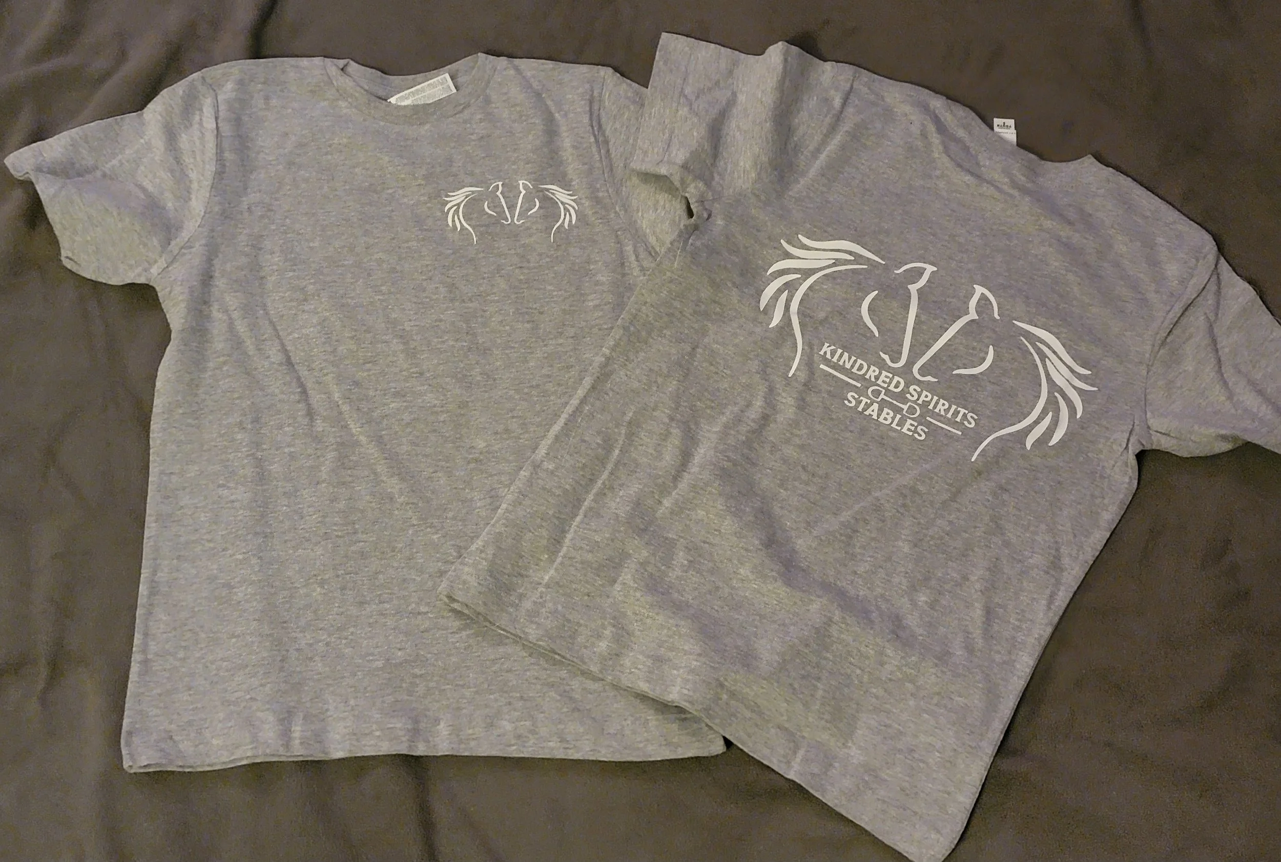 Two gray T-shirts laid out flat on a dark surface. One shirt features a small, minimalist horse head outline at the center. The other shirt displays a larger, decorative horse head with flowing mane and the text "KINDRED SPIRITS STABLES" underneath.