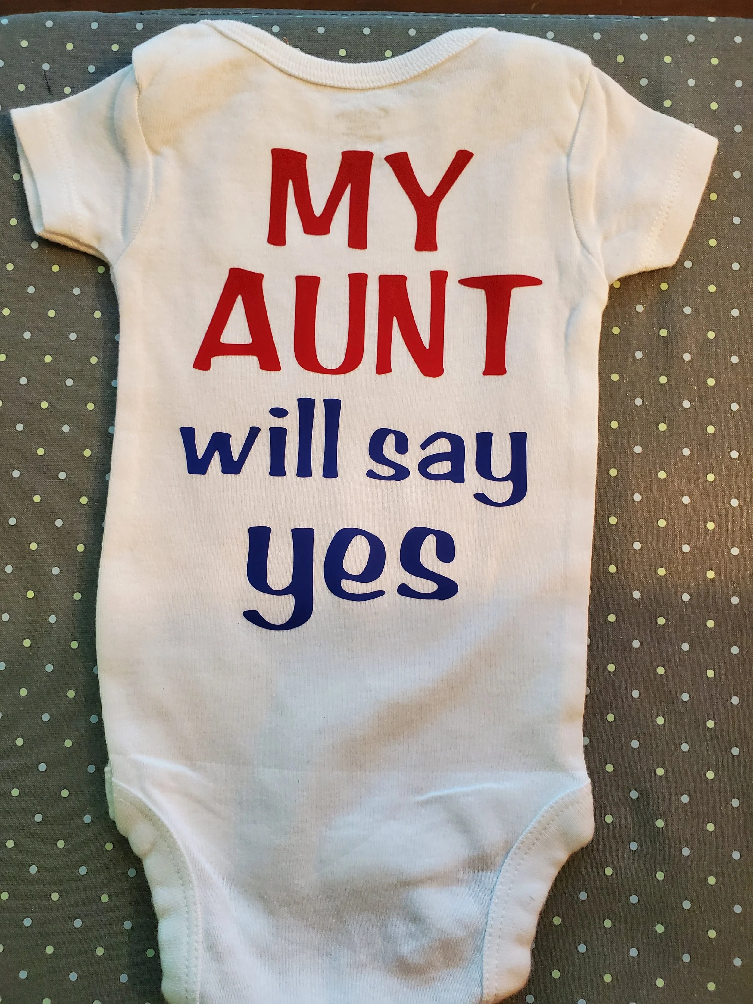 White baby onesie with red and blue text that reads "My Aunt will say yes" on a gray polka-dotted background.