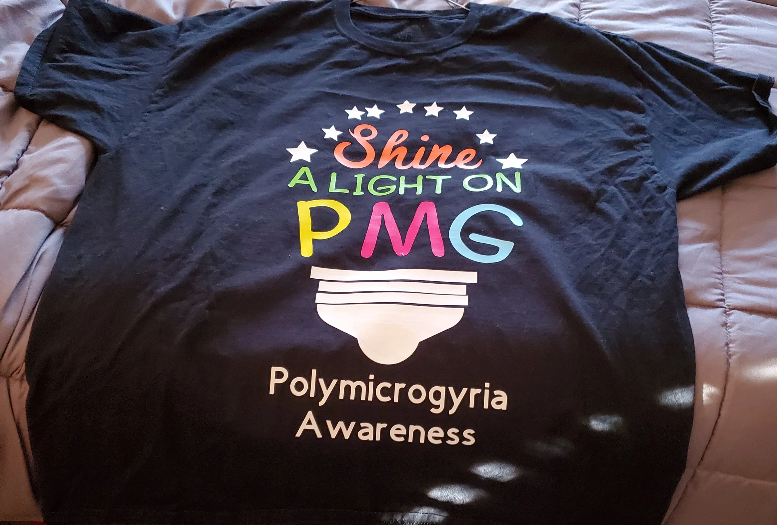 Black T-shirt with colorful text and graphics promoting awareness of Polymicrogyria. The text reads 'Shine a Light on P.M.G.' with a graphic of a uterus and additional text: 'Polymicrogyria Awareness'.