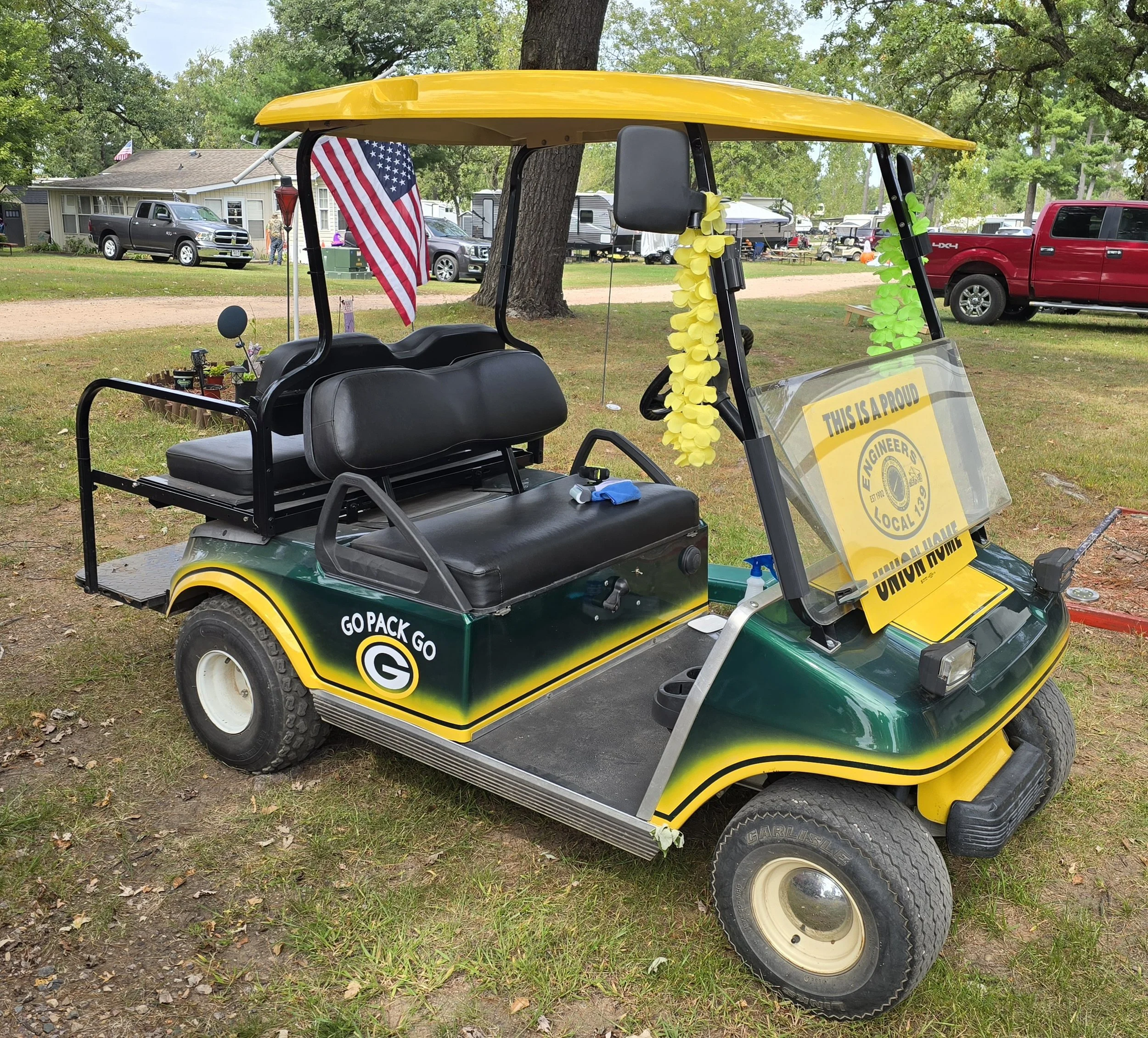 A golf cart decorated with Green Bay Packers colors and logos, with an American flag and yellow flower garlands, parked on grass in a park with trees and cars in the background.