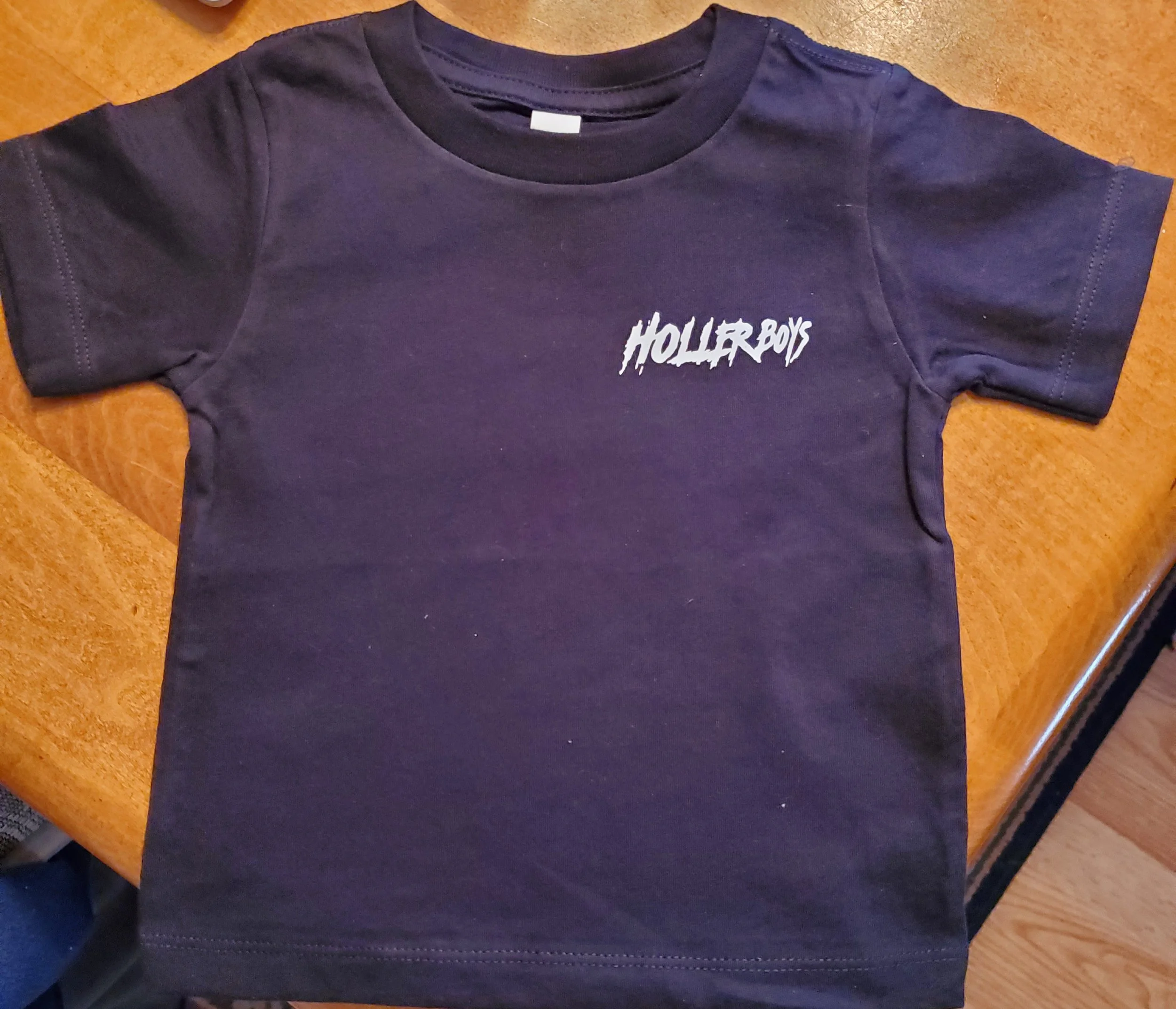 A black Holler Boys t-shirt with white text logo on the upper left side, laid flat on a wooden surface.