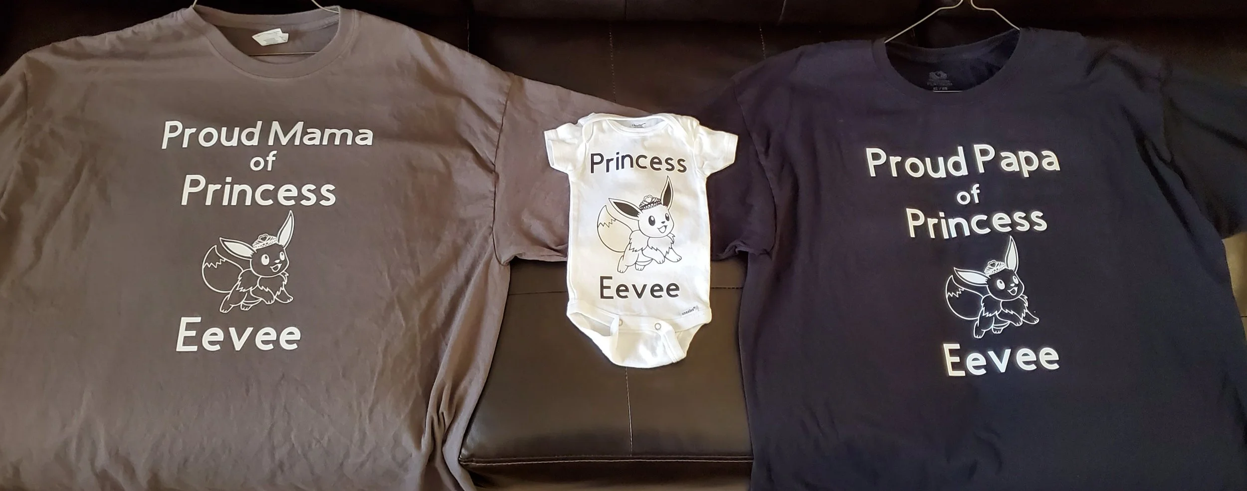Three shirts with Pokémon Eevee themed designs. The left shirt is gray with the text 'Proud Mama of Princess Eevee' and an illustration of Eevee. The middle shirt is a white onesie with the text 'Princess Eevee' and an illustration of Eevee. The righ
