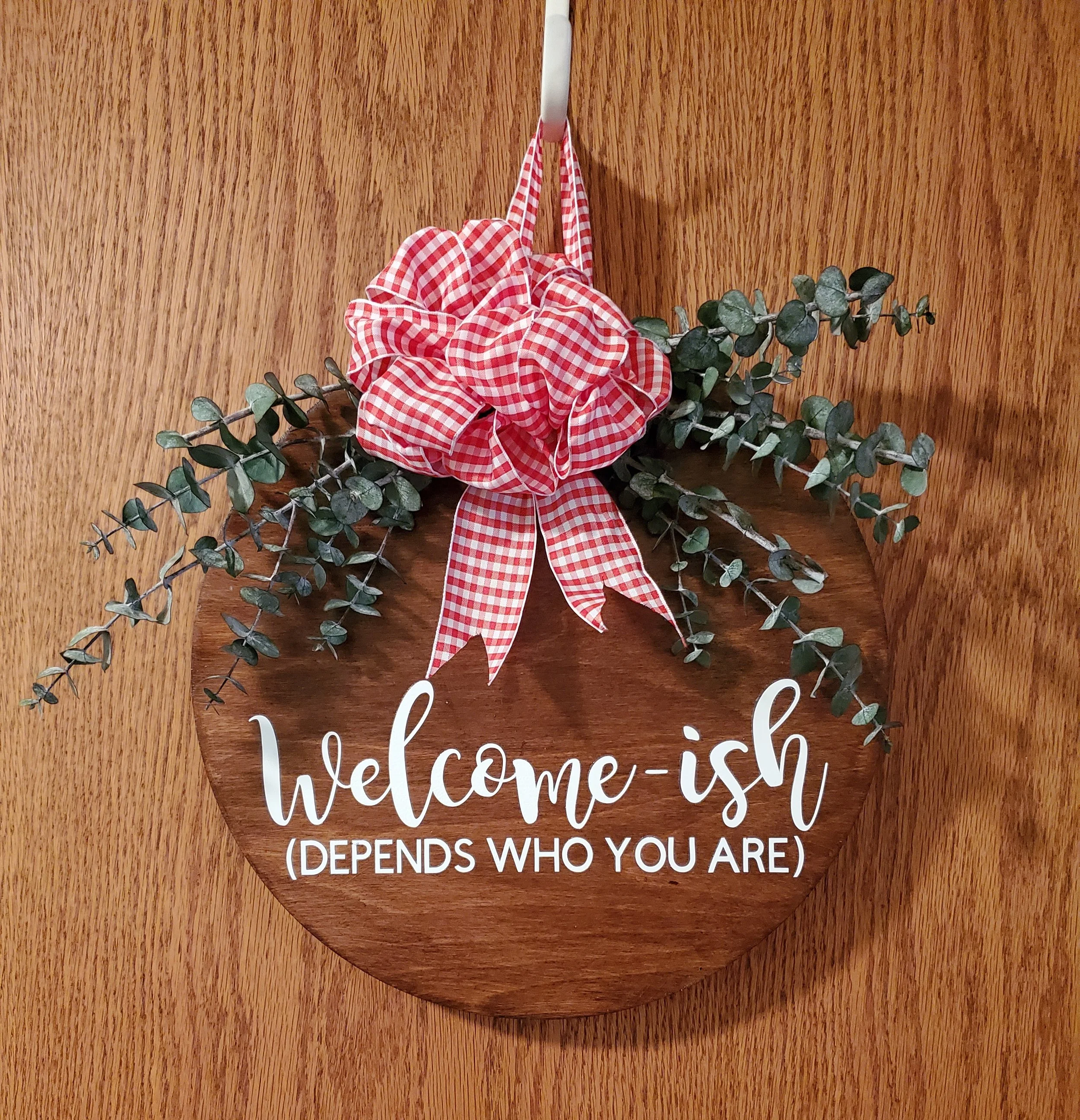 Decorative wooden sign with white text that reads 'welcome-ish (DEPENDS WHO YOU ARE)' with a red gingham bow and eucalyptus leaves attached, hanging on a wooden door.