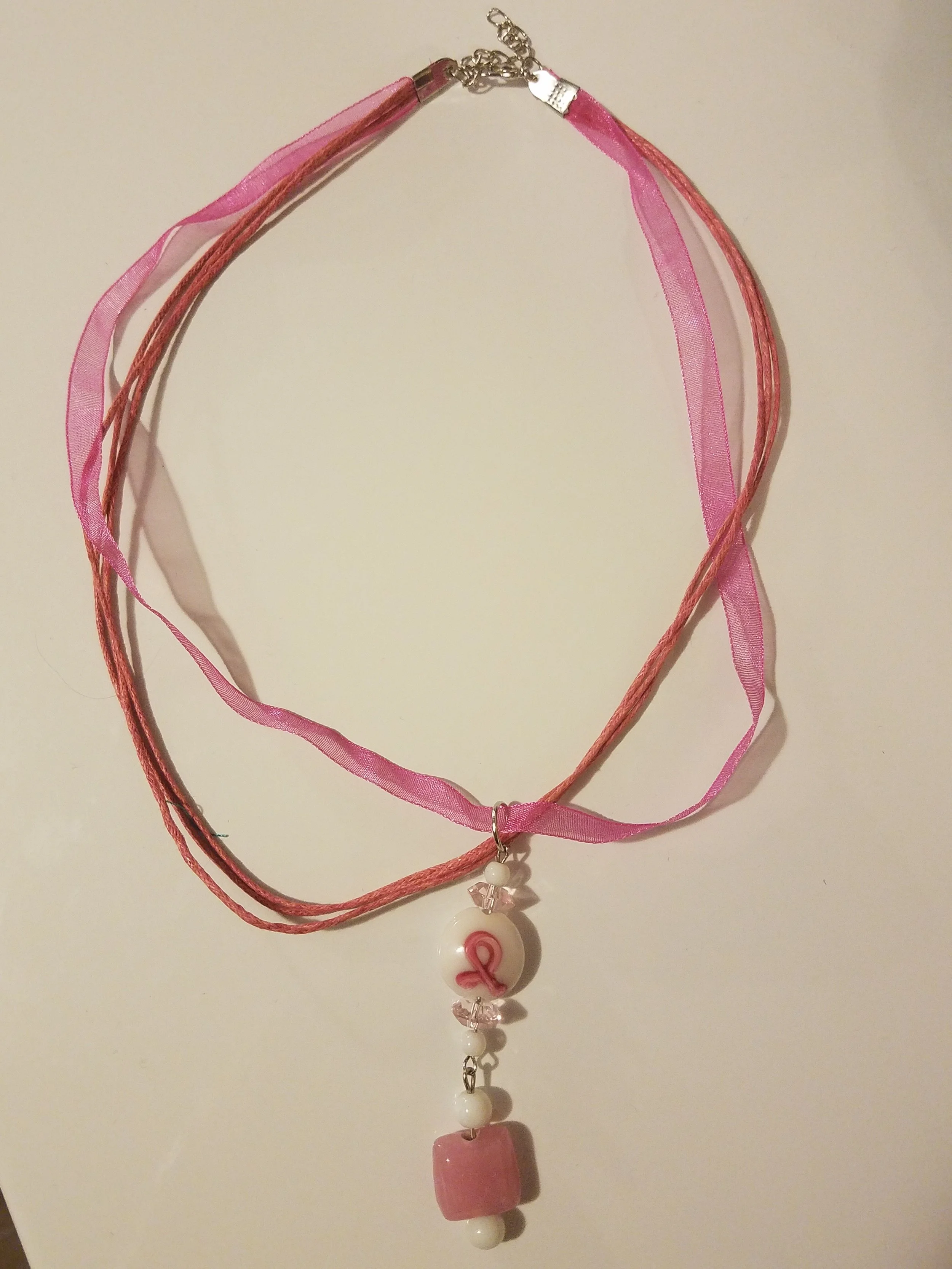 A pink ribbon necklace with a pink ribbon charm, pink beads, and a pink cube pendant.