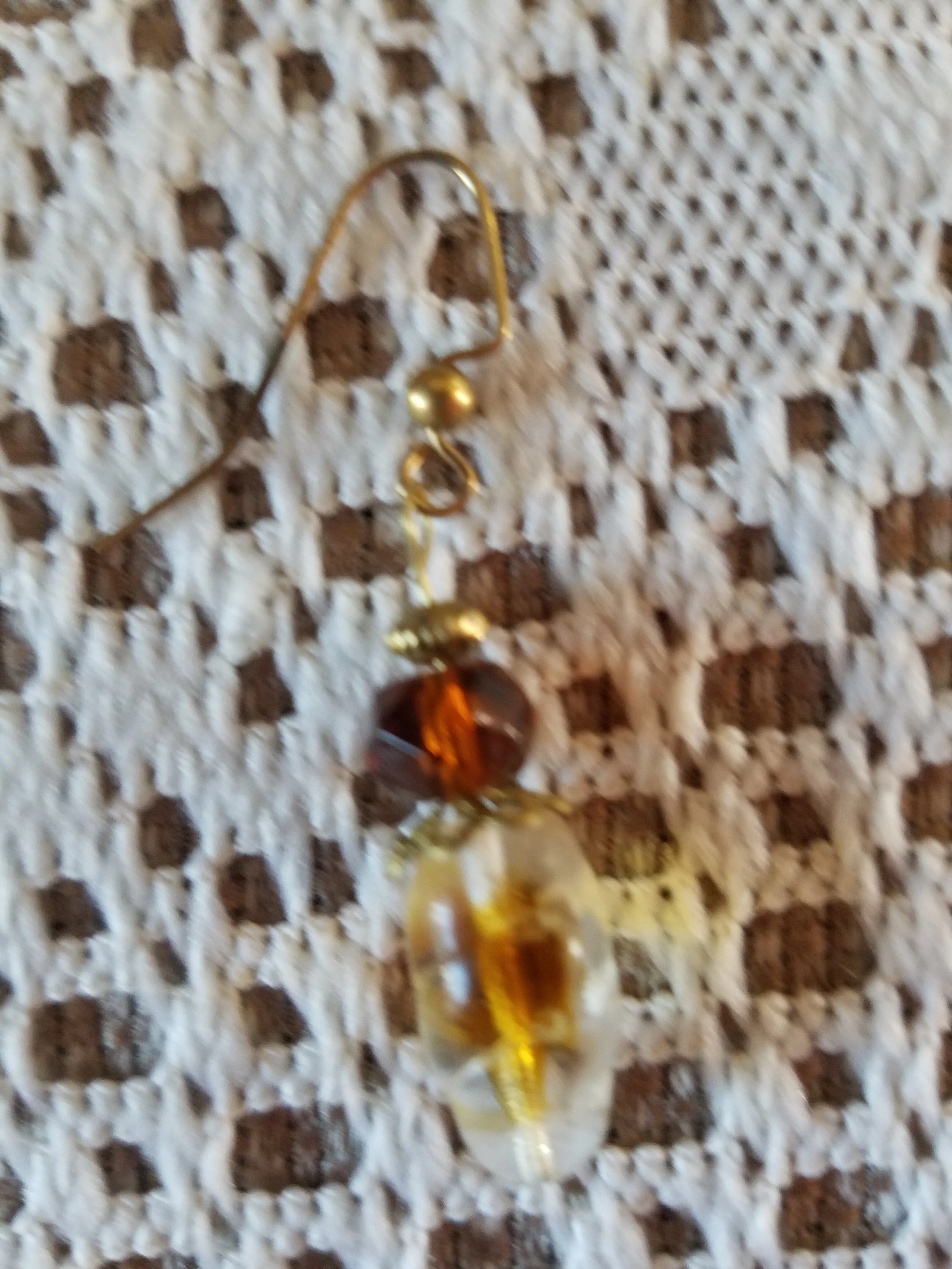 A decorative earring made of gold-colored wire, a large amber-colored bead, a smaller dark amber bead, and a pearl, placed on a textured beige and brown surface.