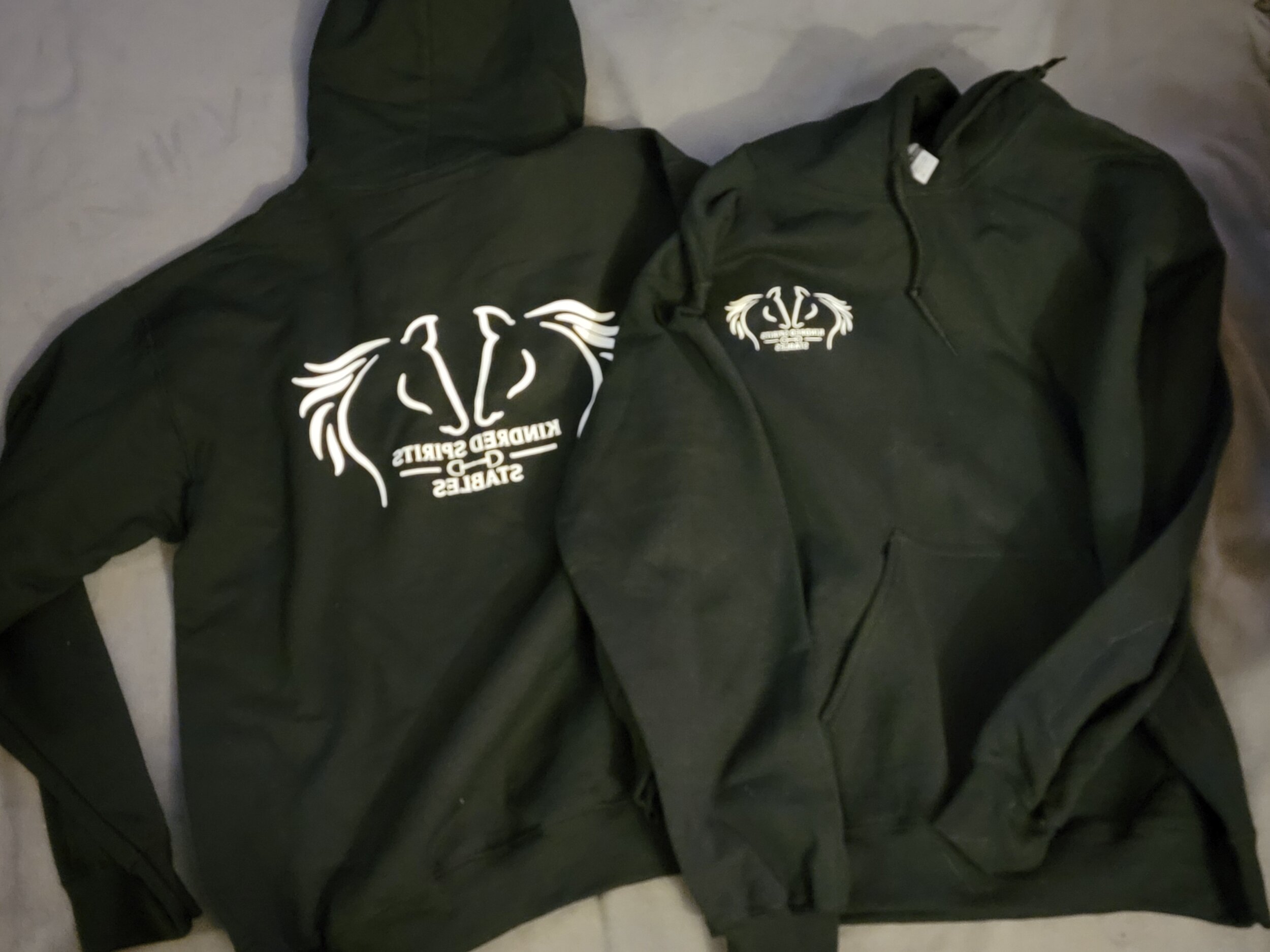 Two black hooded sweatshirts with white printed logos and text, laid out on a gray surface. One sweatshirt has a large logo on the back, and the other has a smaller logo on the left chest area.