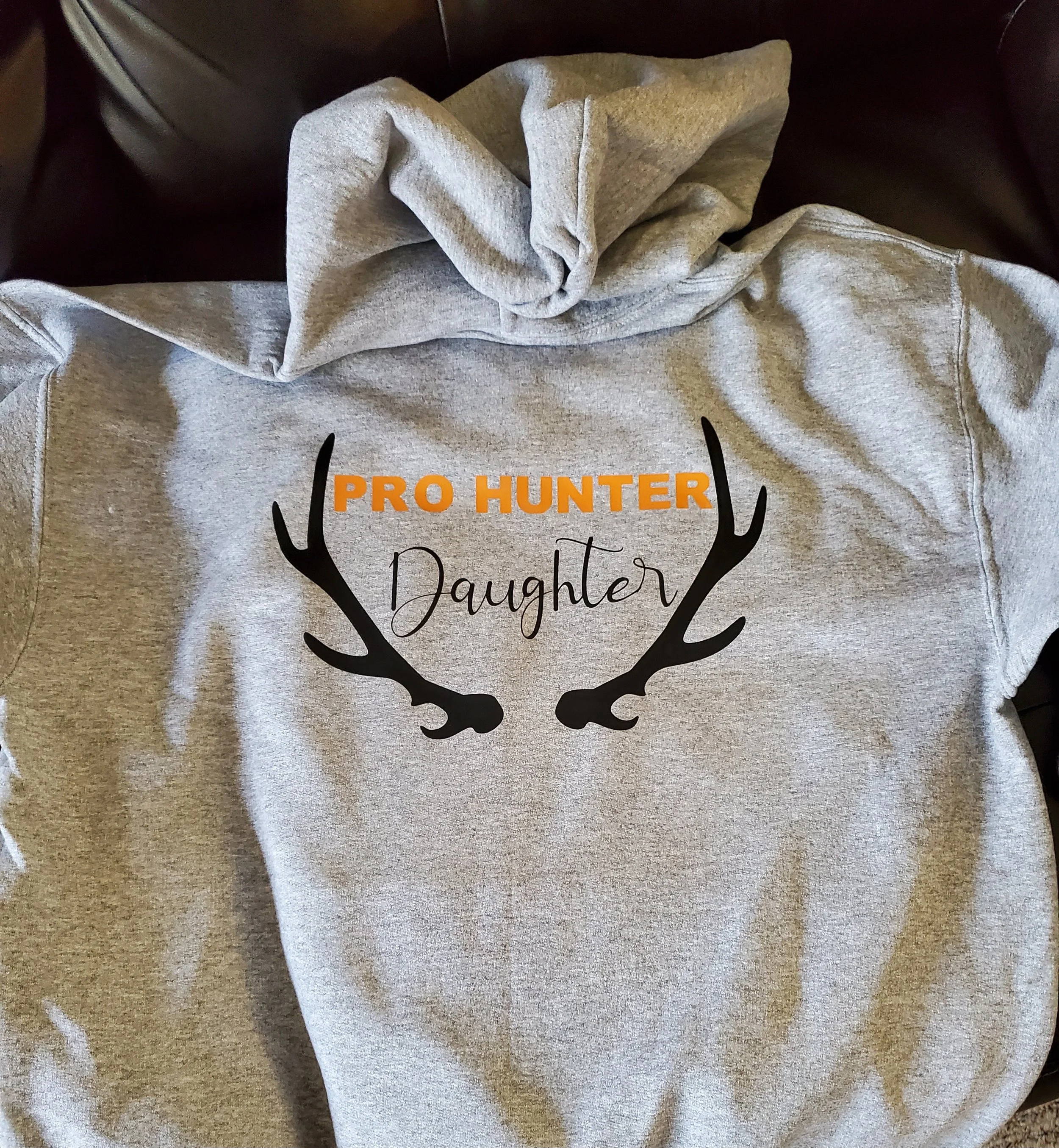 Gray hoodie with black antlers and text that reads "Pro Hunter Daughter" in orange and black, placed on a surface.