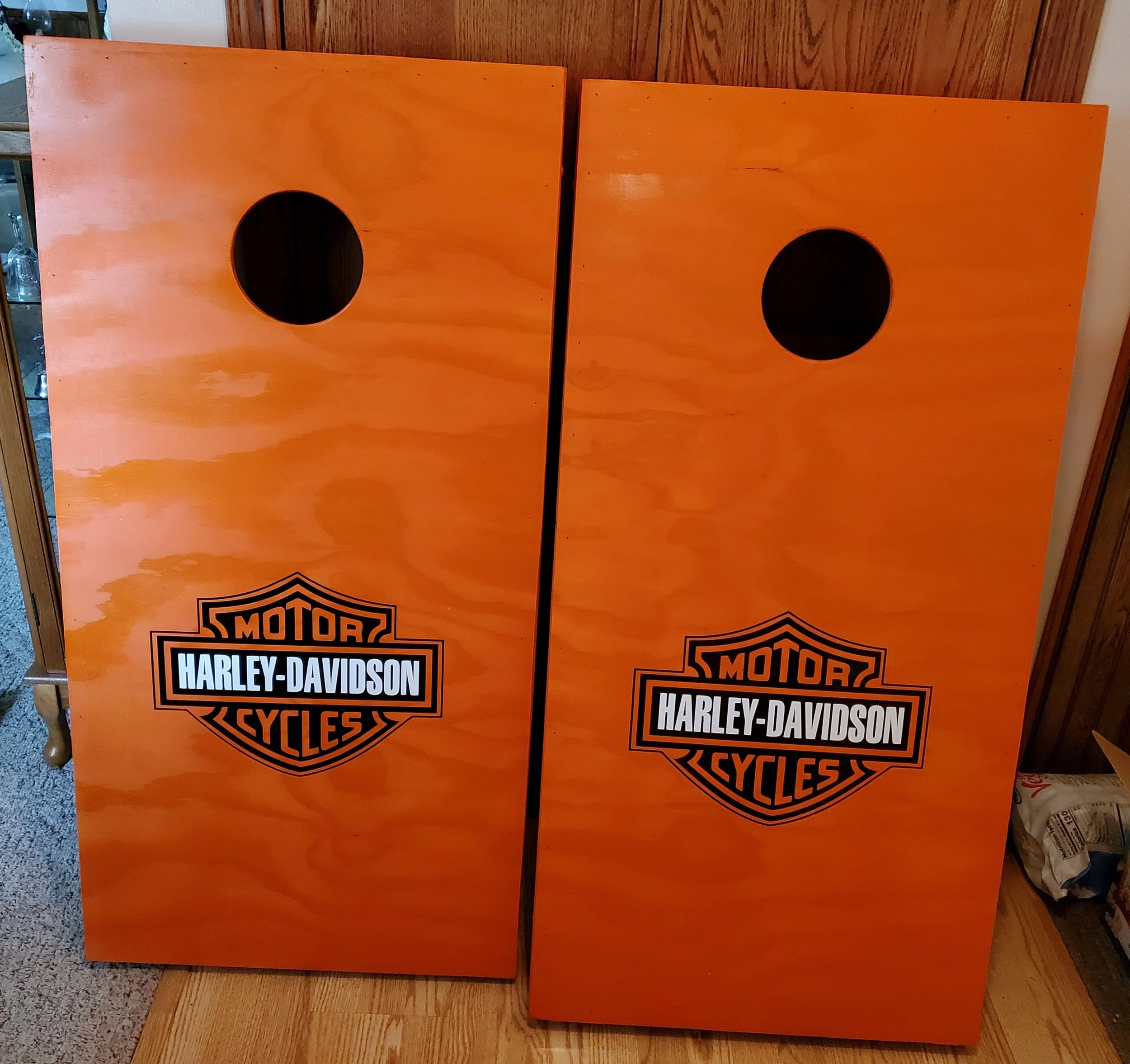 Two wooden Harley-Davidson branded cornhole game boards with circular hole near the top, leaning against a wood-paneled wall.