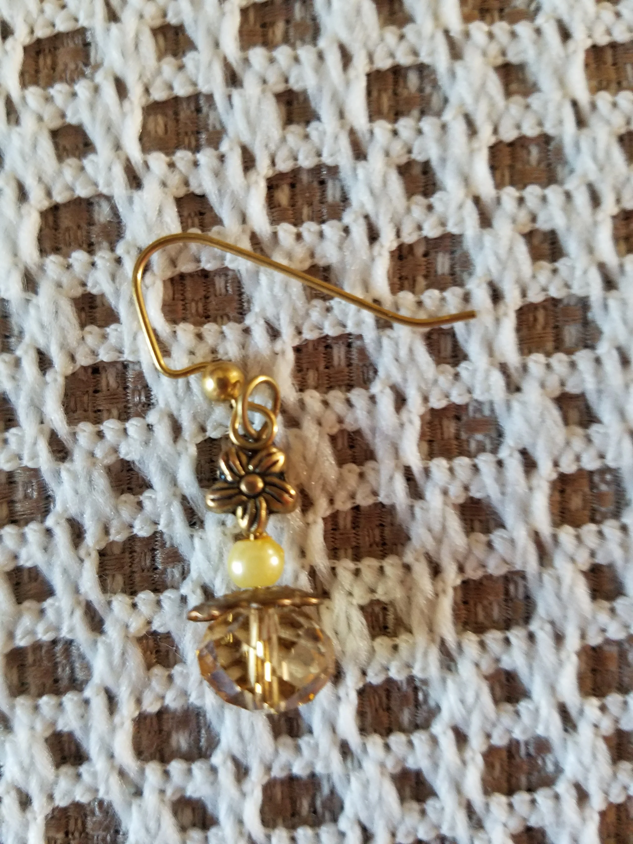 A gold fishhook earring with a flower, pearl, and faceted crystal charm on a white textured fabric background.