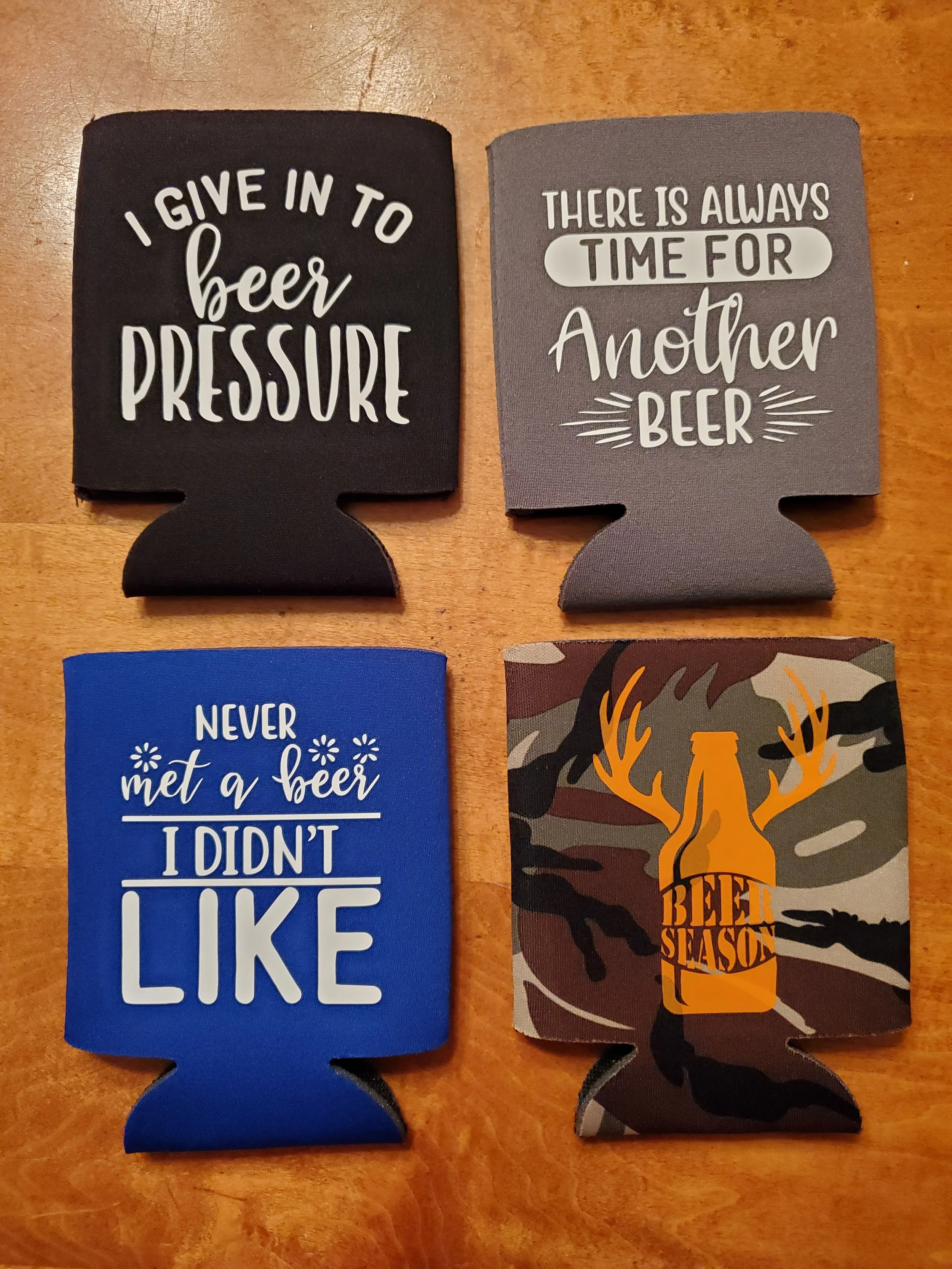 Four beer-themed coasters with humorous sayings, placed on a wooden surface. The sayings include 'I give in to beer pressure', 'There is always time for another beer', 'Never met a beer I didn't like', and a camouflage background with a beer bottle a