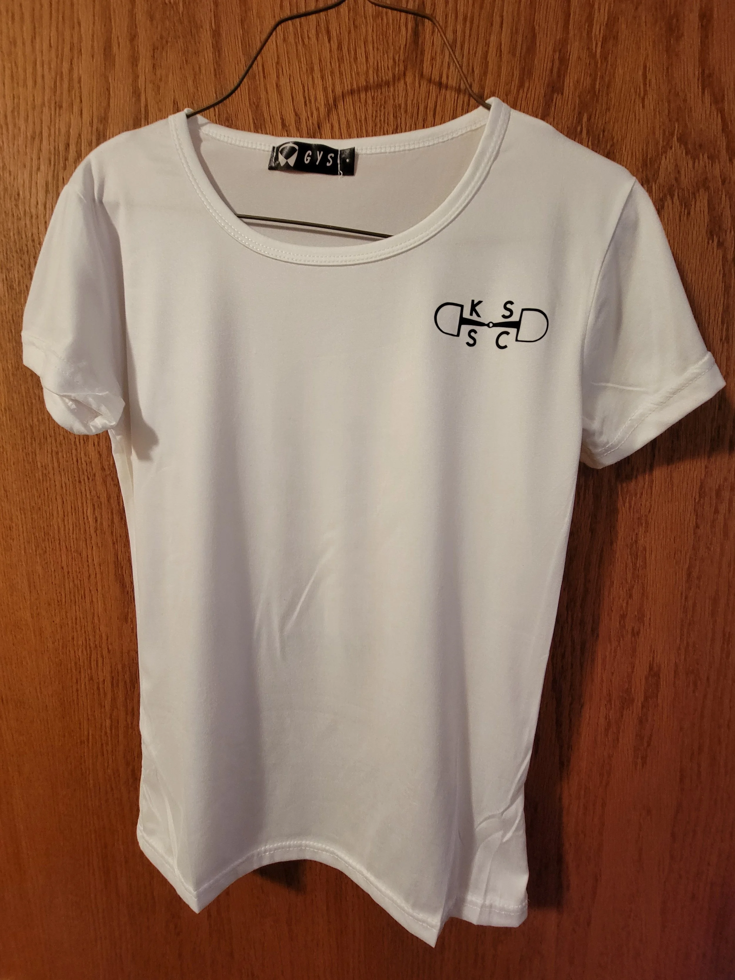 White T-shirt hanging on a metal hanger against a wooden background. The T-shirt has a small black and white graphic of a medicine capsule and the letters K, S, C, and S connected by lines on the front right side.