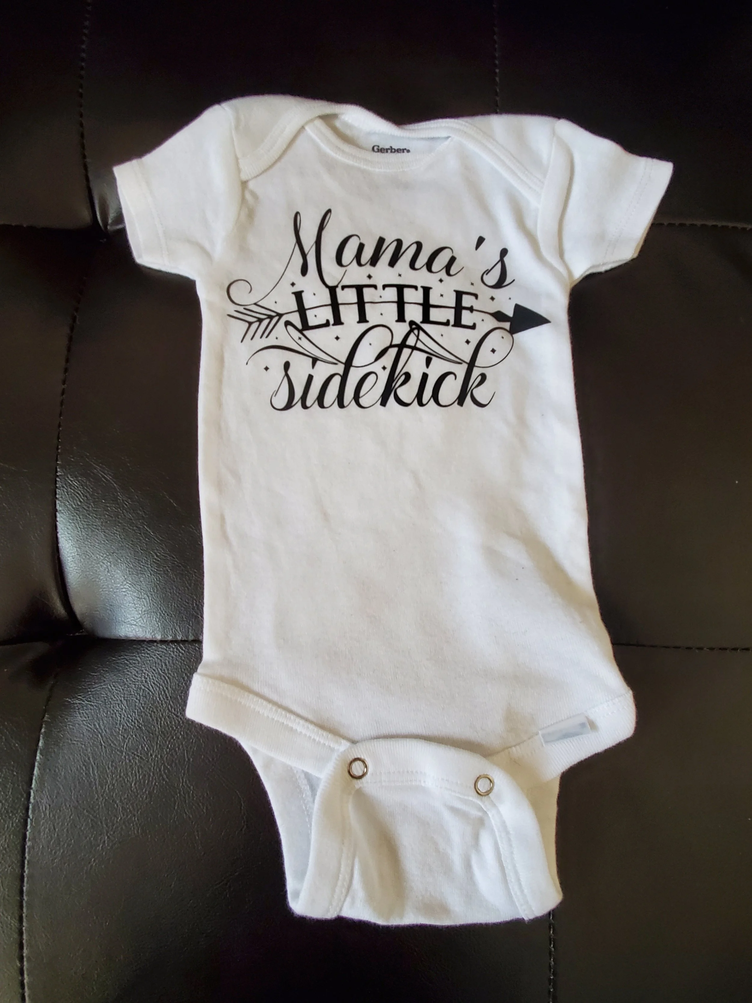 White baby onesie with black text that reads 'Mama's Little Sidekick' and an arrow design.
