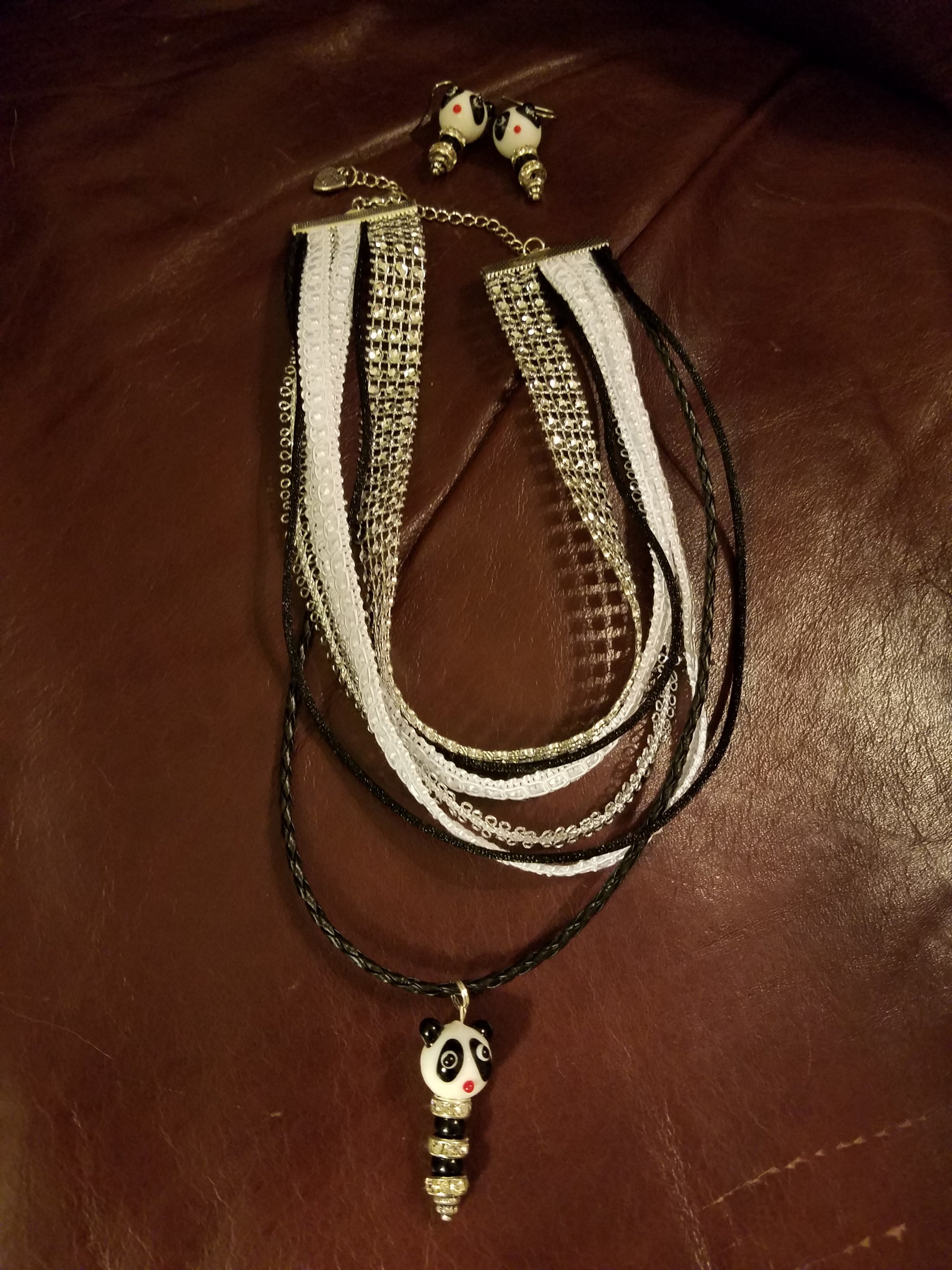 A panda-shaped necklace with a black cord and silver beads, and a pair of panda-shaped earrings with white, black, and red details placed on a brown leather surface.