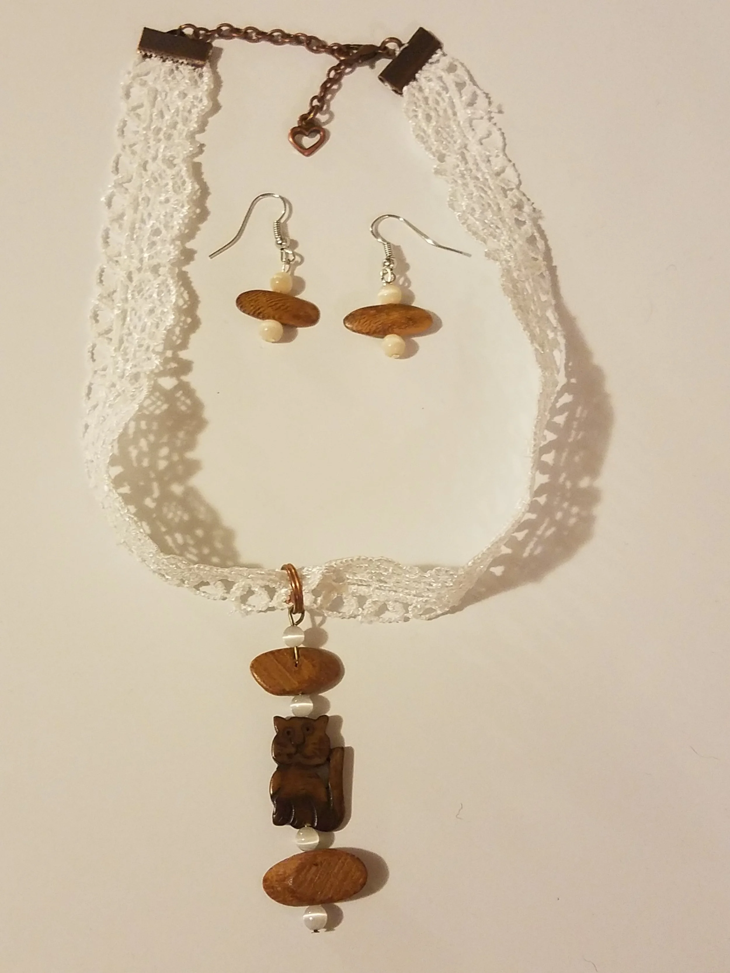 A set of jewelry including a lace choker necklace with a heart-shaped chain clasp, a pair of wooden and bead earrings, and a matching wooden and bead pendant necklace with a carved animal figure, all hanging against a light background.