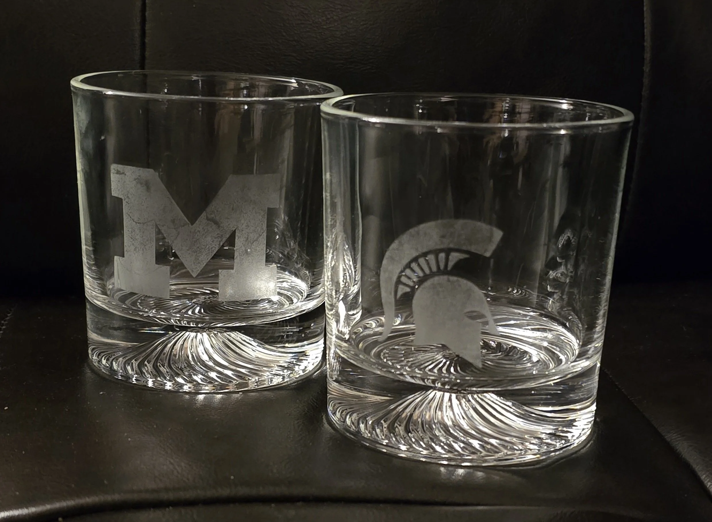 Two clear tumblers with etched monogram and Spartan helmet logos, filled with water, placed on a dark surface.