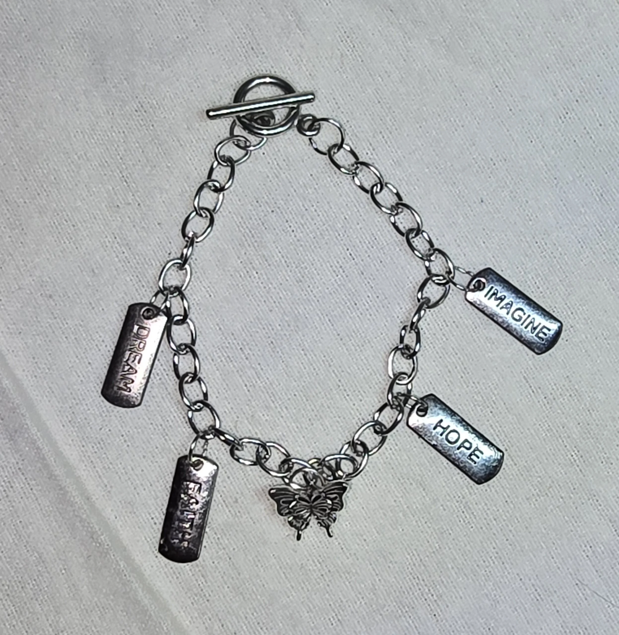 A silver charm bracelet with four rectangular charms engraved with words and a butterfly charm. The words on the charms are 'DREAM,' 'IMAGINE,' 'HOPE,' and 'DREAM' again.