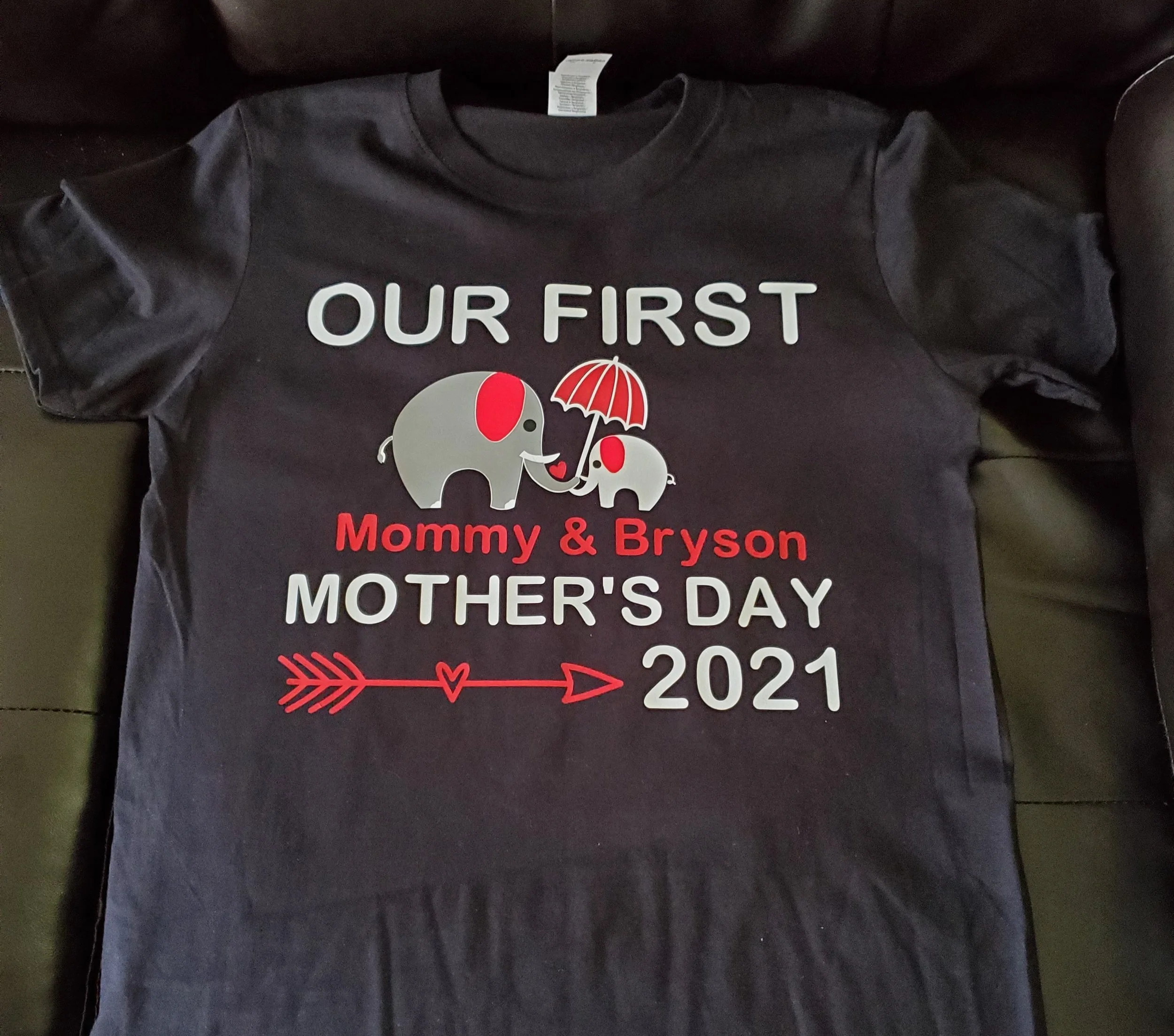 Black T-shirt with a graphic of two gray elephants, one larger and one smaller, holding a red umbrella. Text reads "Our First Mommy & Bryson Mother's Day 2021" with a red arrow and heart design.
