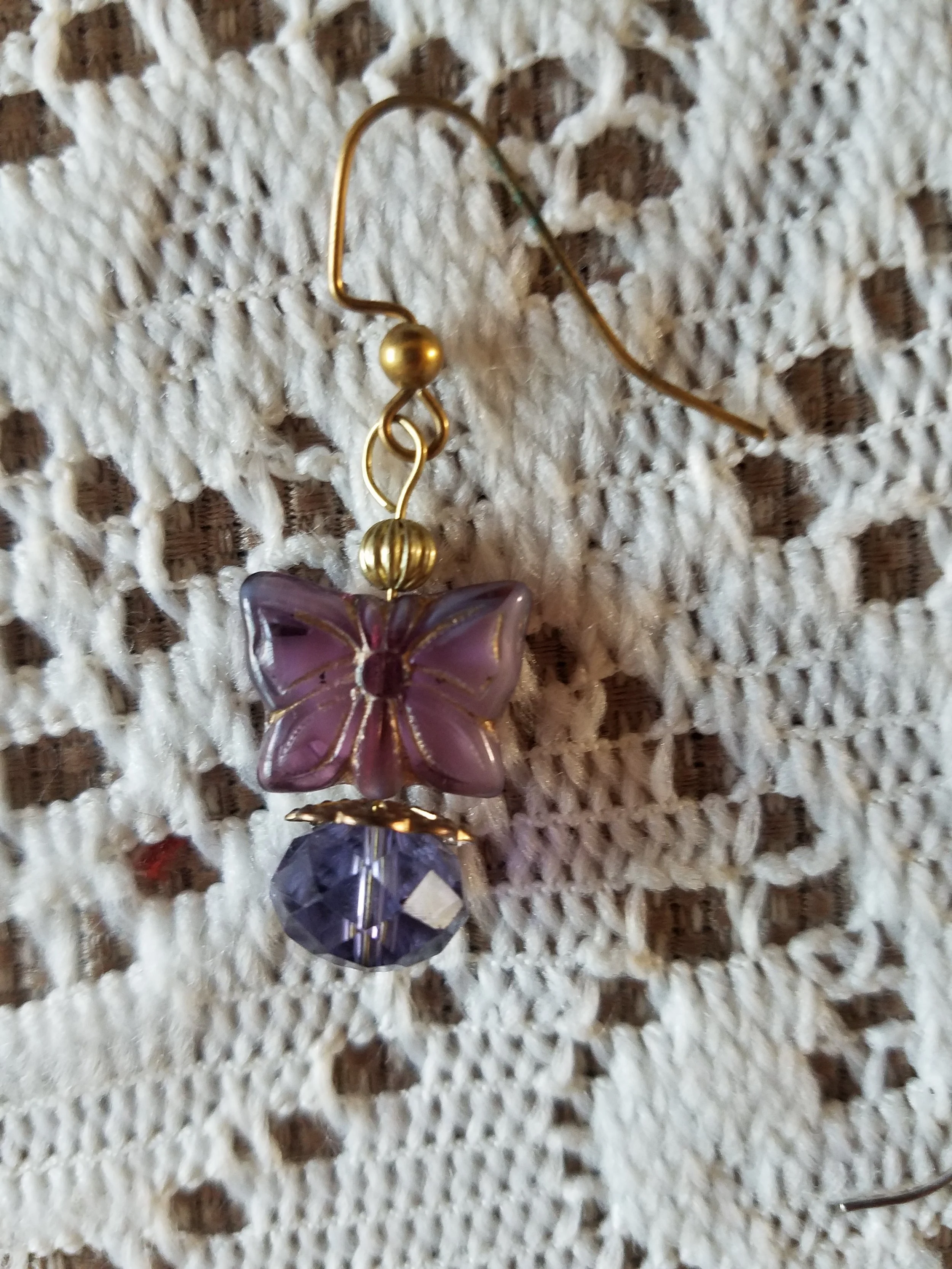 Earring with a purple butterfly pendant and a purple faceted bead hanging below, on a white crocheted background.