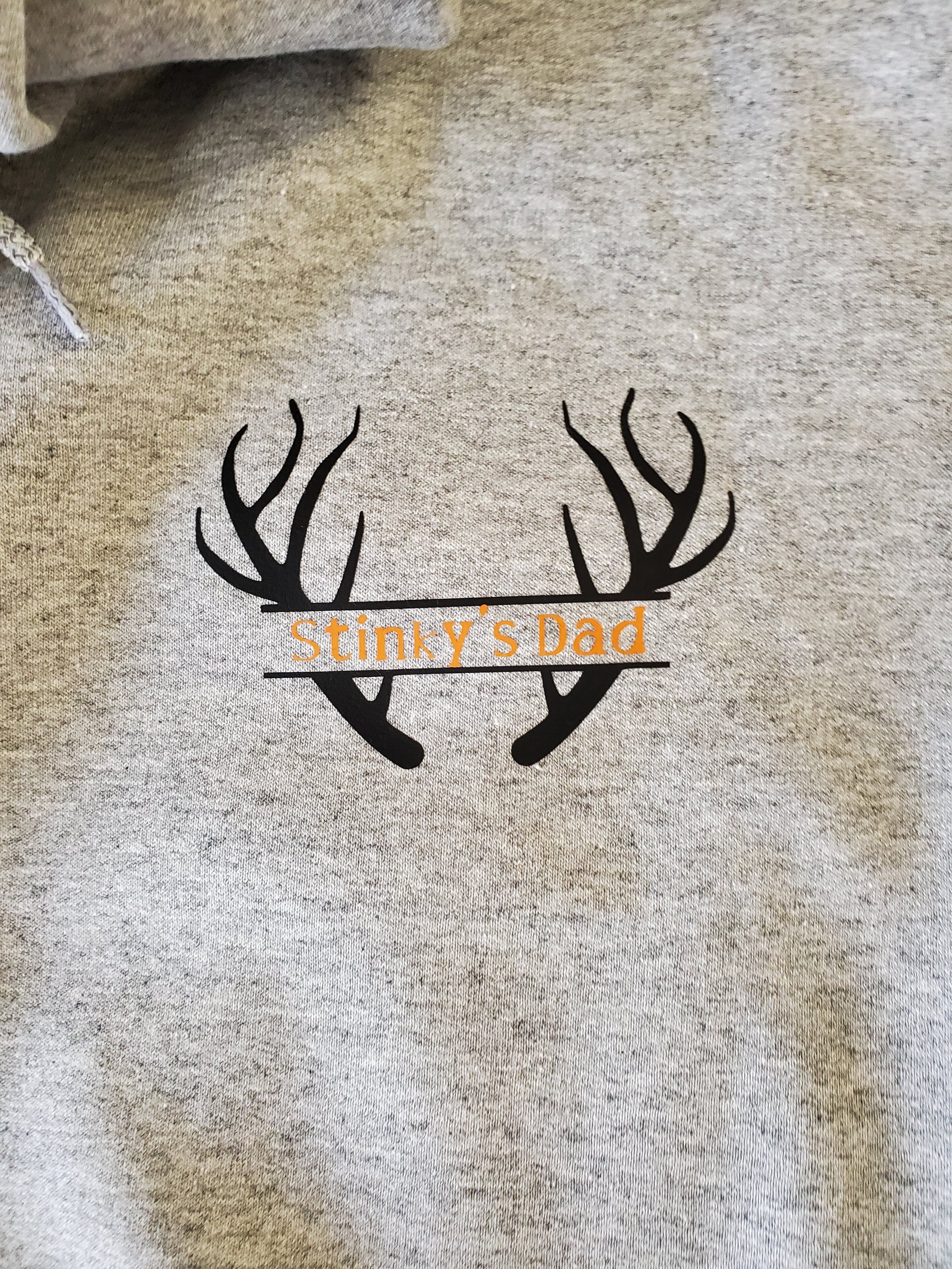 Graphic design of black deer antlers with the text 'Stinky's Dad' in orange across the middle on a gray fabric background.