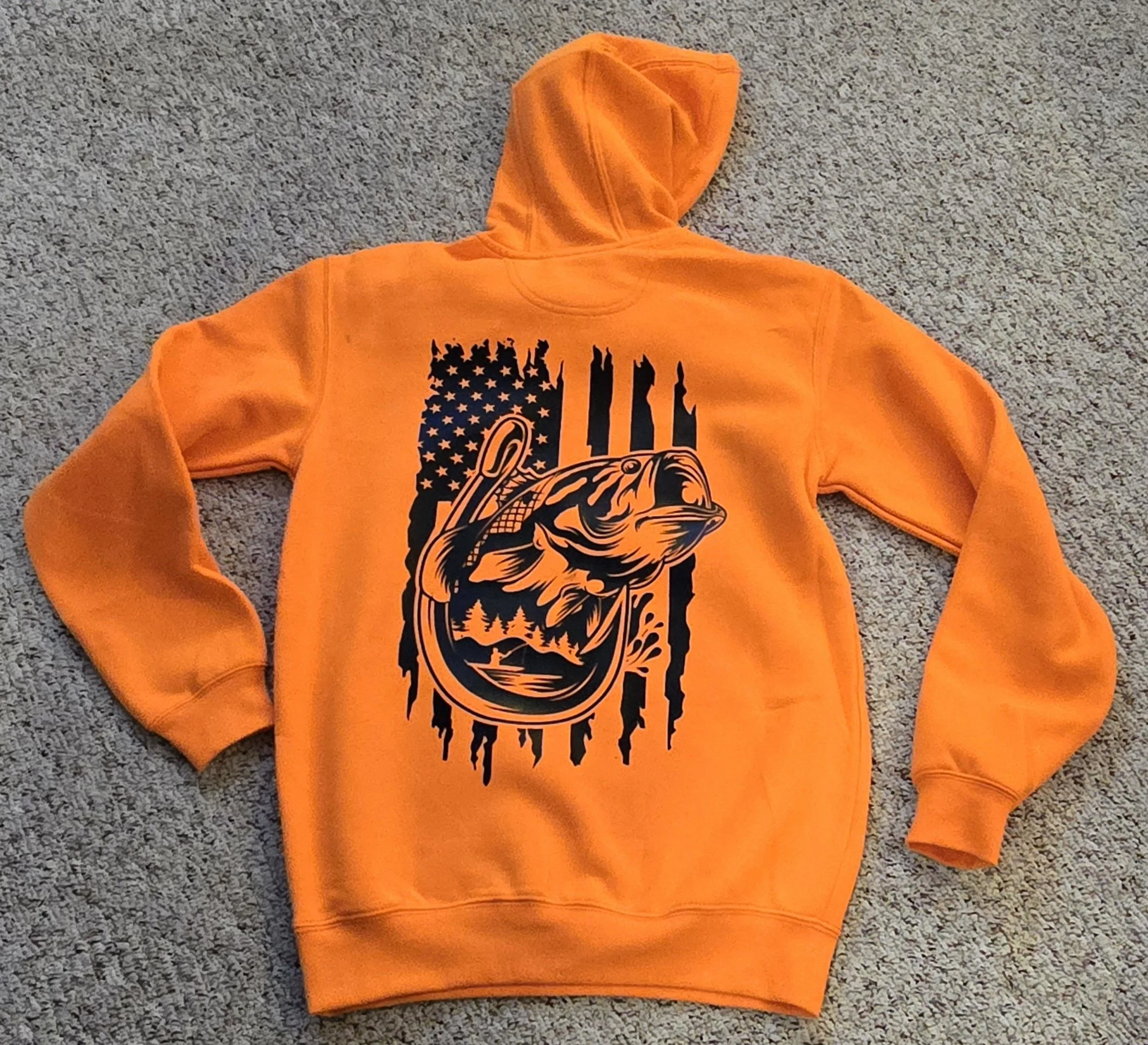 Orange hoodie with a black graphic of a fish and an American flag design on the back, laid flat on a carpeted floor.