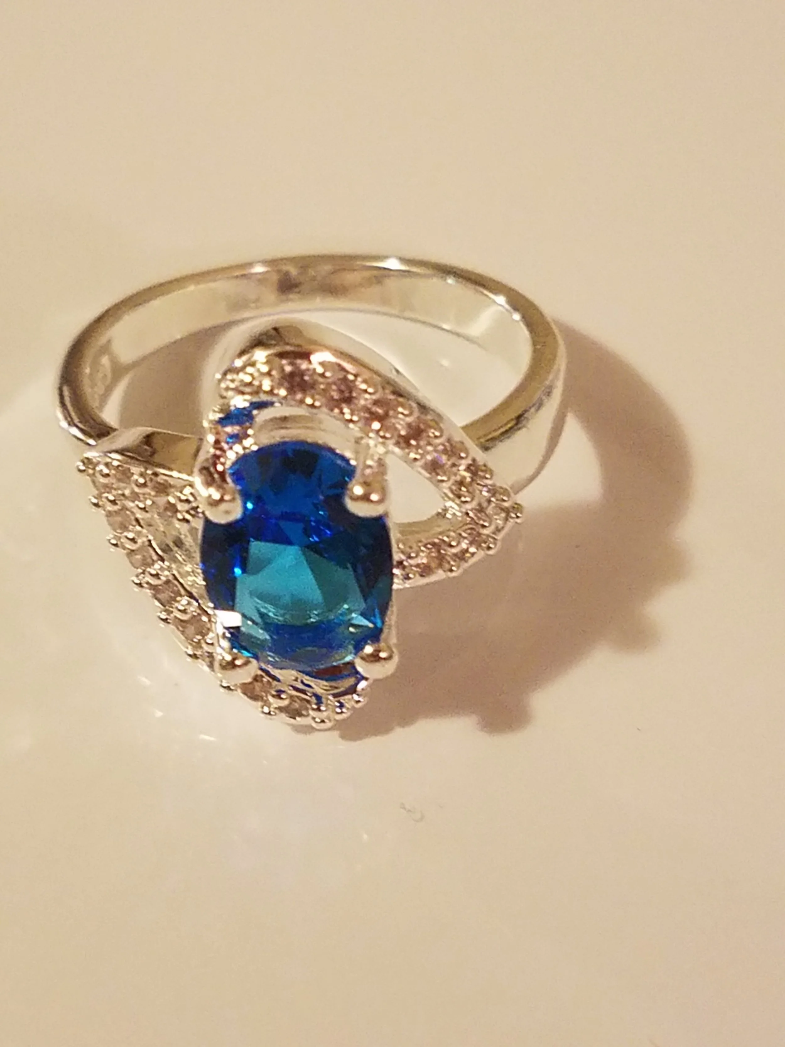 Silver ring with a large oval blue gemstone in the center, surrounded by small clear stones, set against a plain white background.