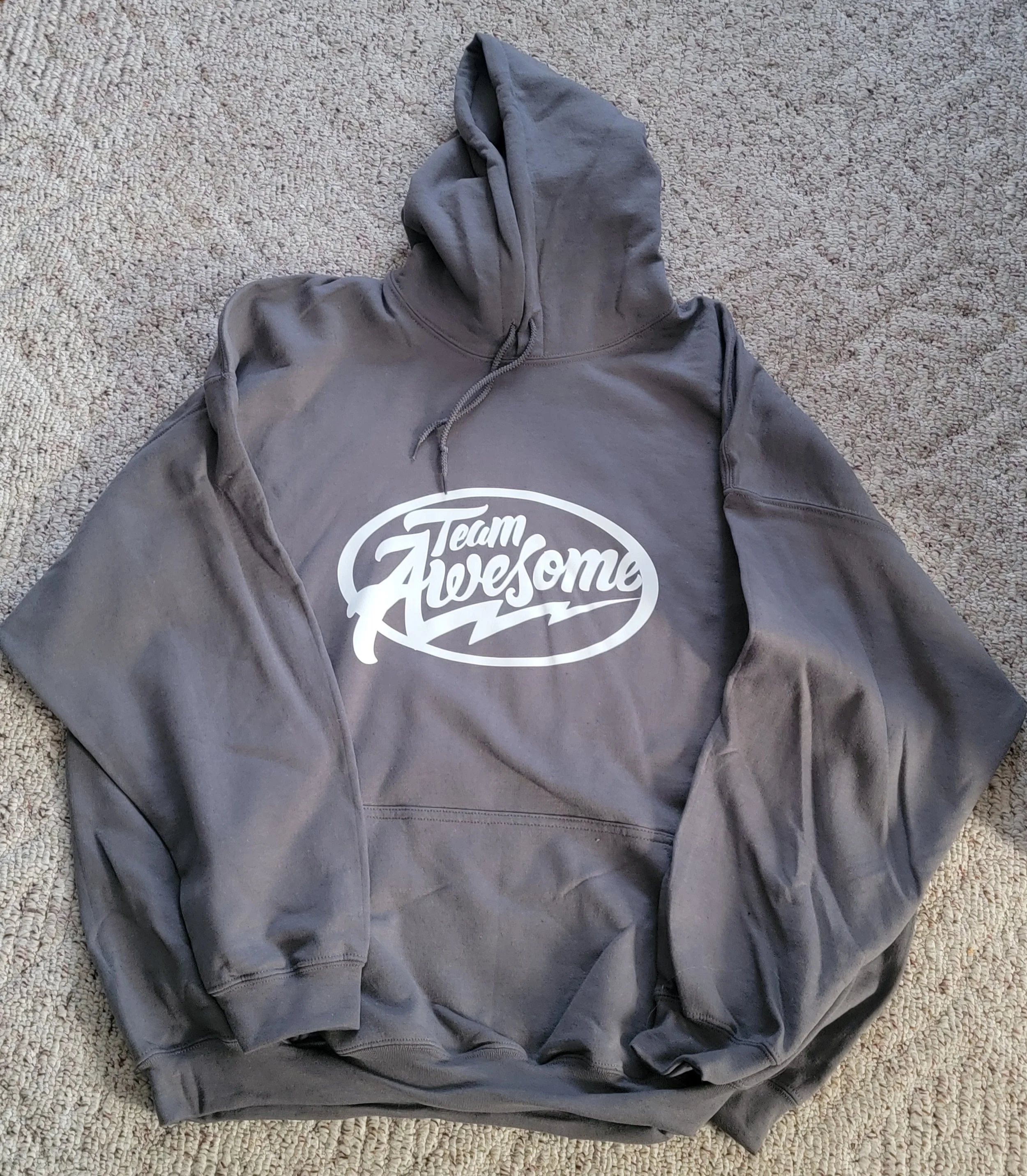 Gray hoodie with a white print that says 'Team Awesome' on the front, laid flat on a beige carpet.