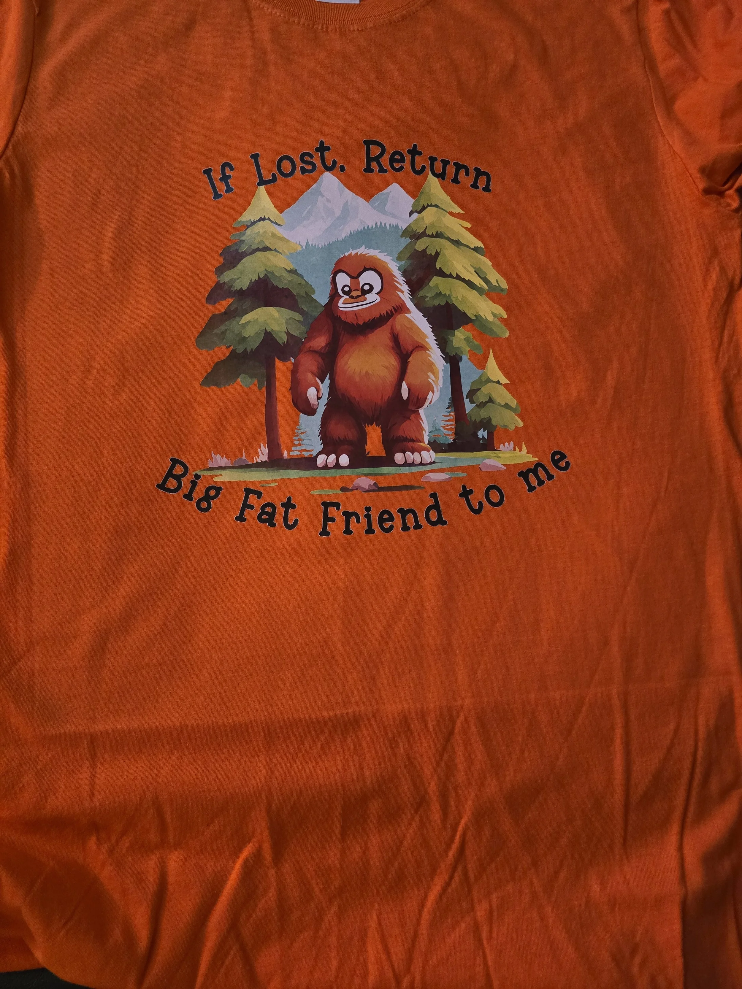 Orange T-shirt with a cartoon graphic of a Bigfoot surrounded by trees, mountains, and a river. The text reads: 'If Lost, Return Big Fat Friend to me'.