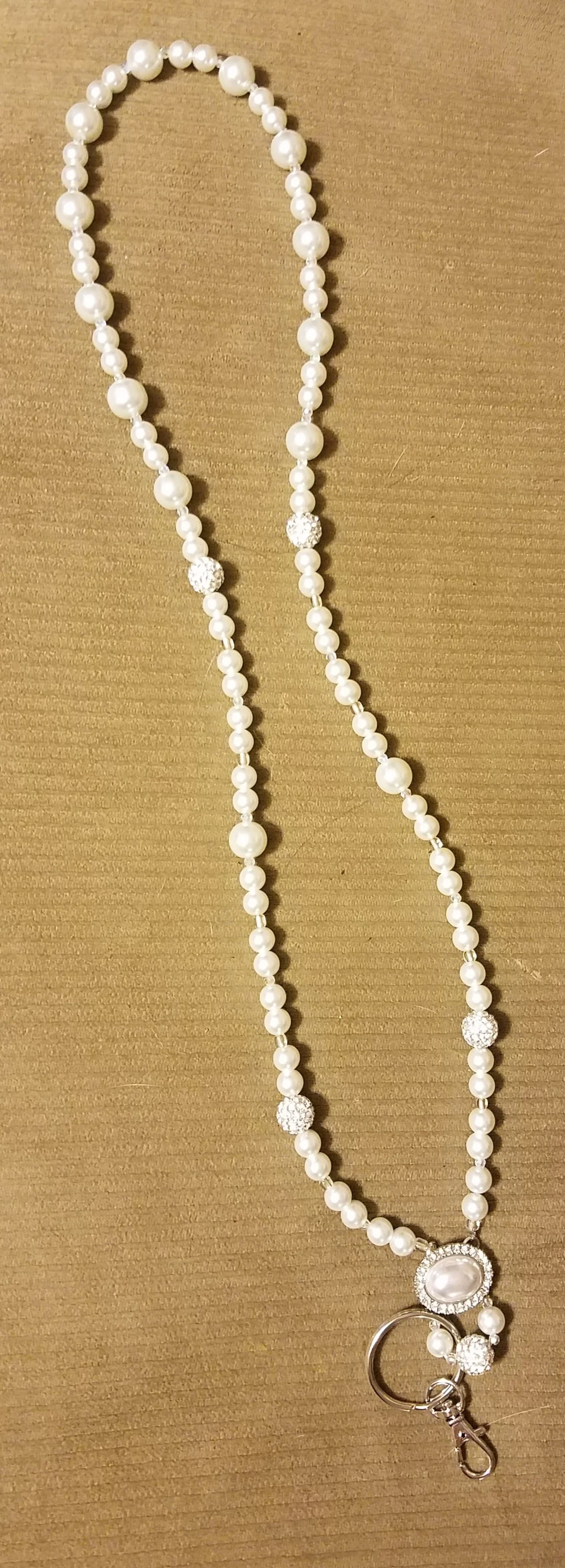 A pearl necklace with a keychain pendant, featuring white pearls, crystal beads, and a large pearl centerpiece with rhinestones on a brown surface.