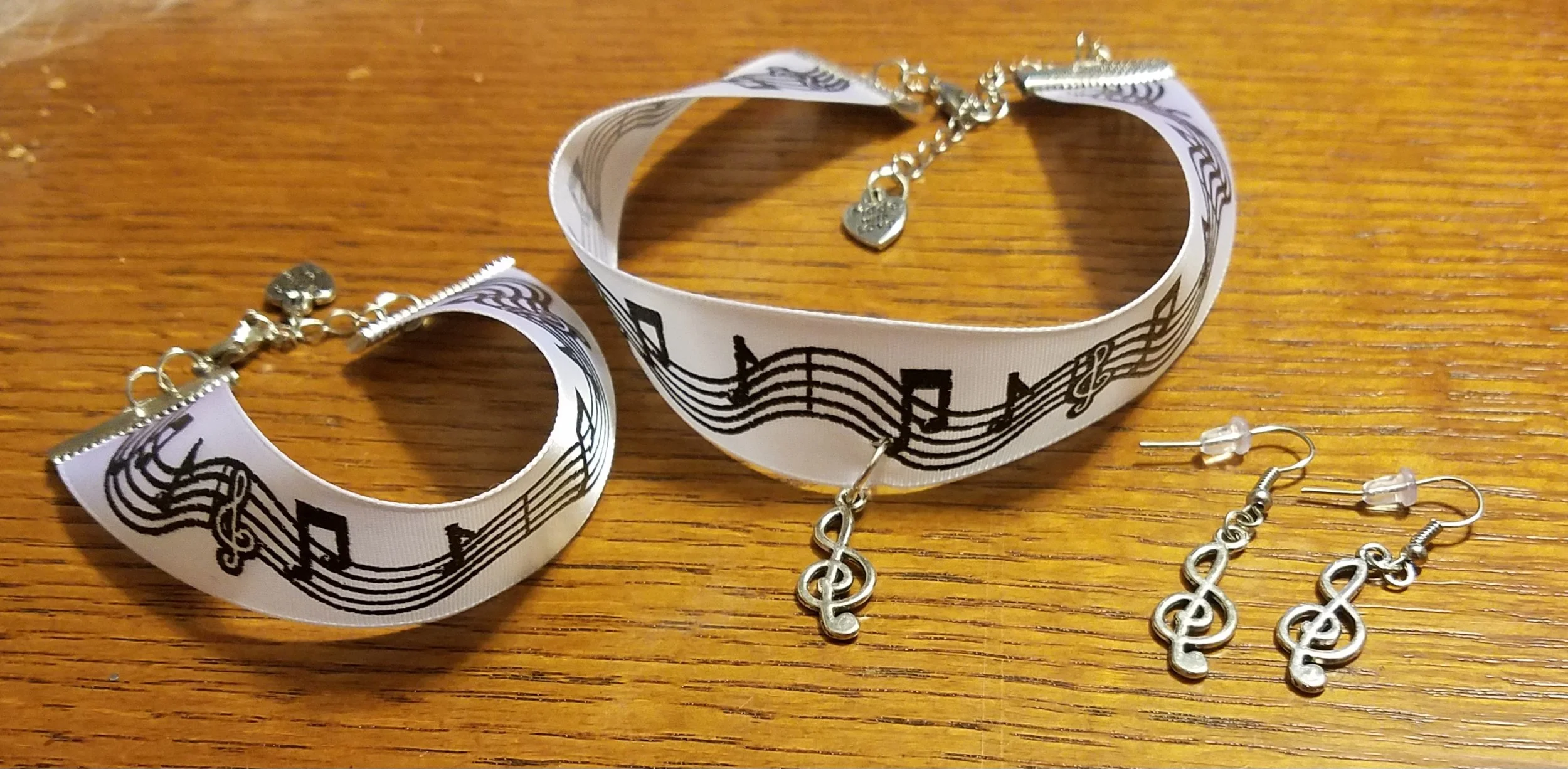 Music-themed jewelry set including two ribbon-style bracelets with musical note and staff designs, and matching earrings with treble clef charms, all displayed on a wooden surface.