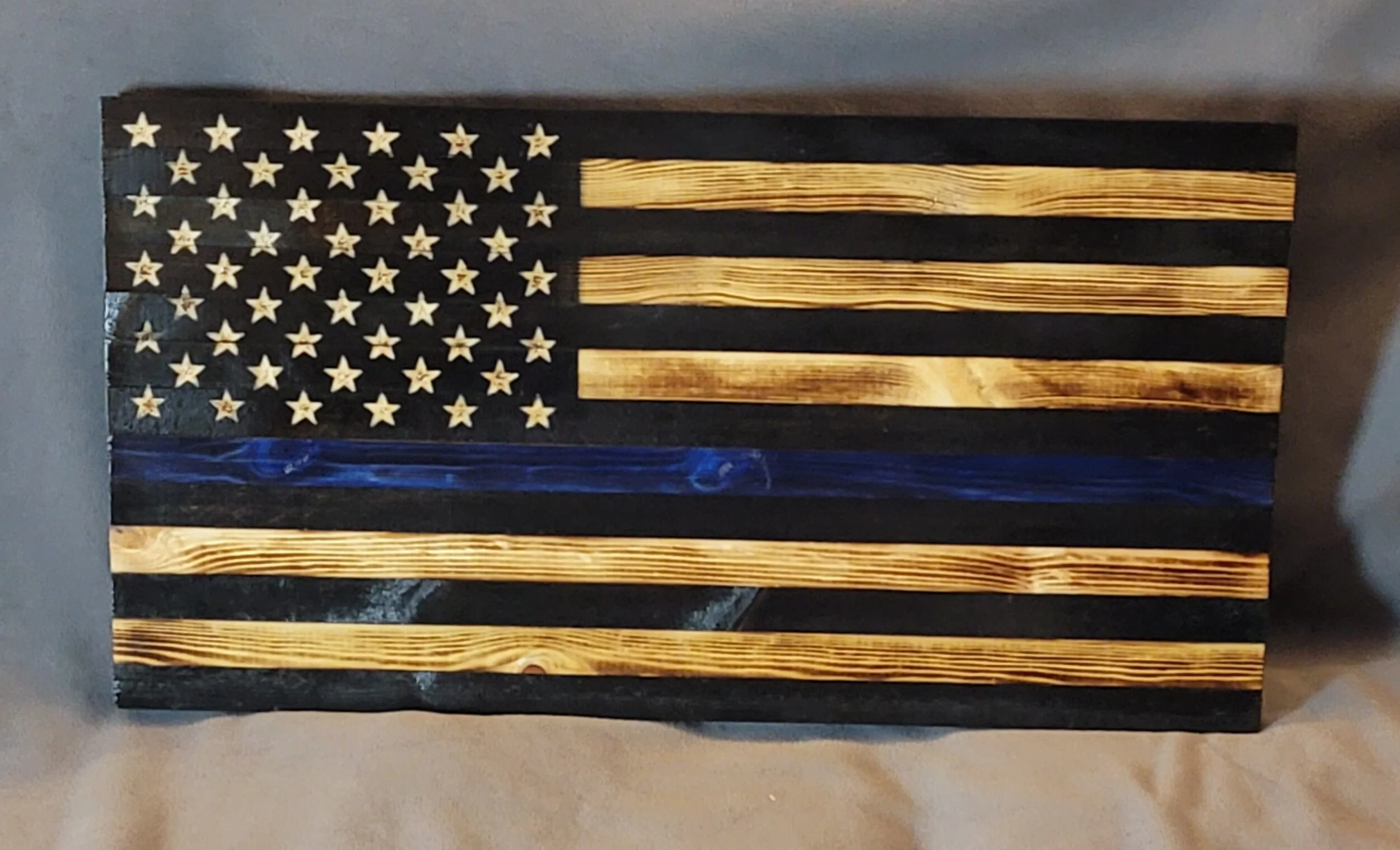Wooden American flag with black and natural wood stripes, and a blue line, mounted on a wall.