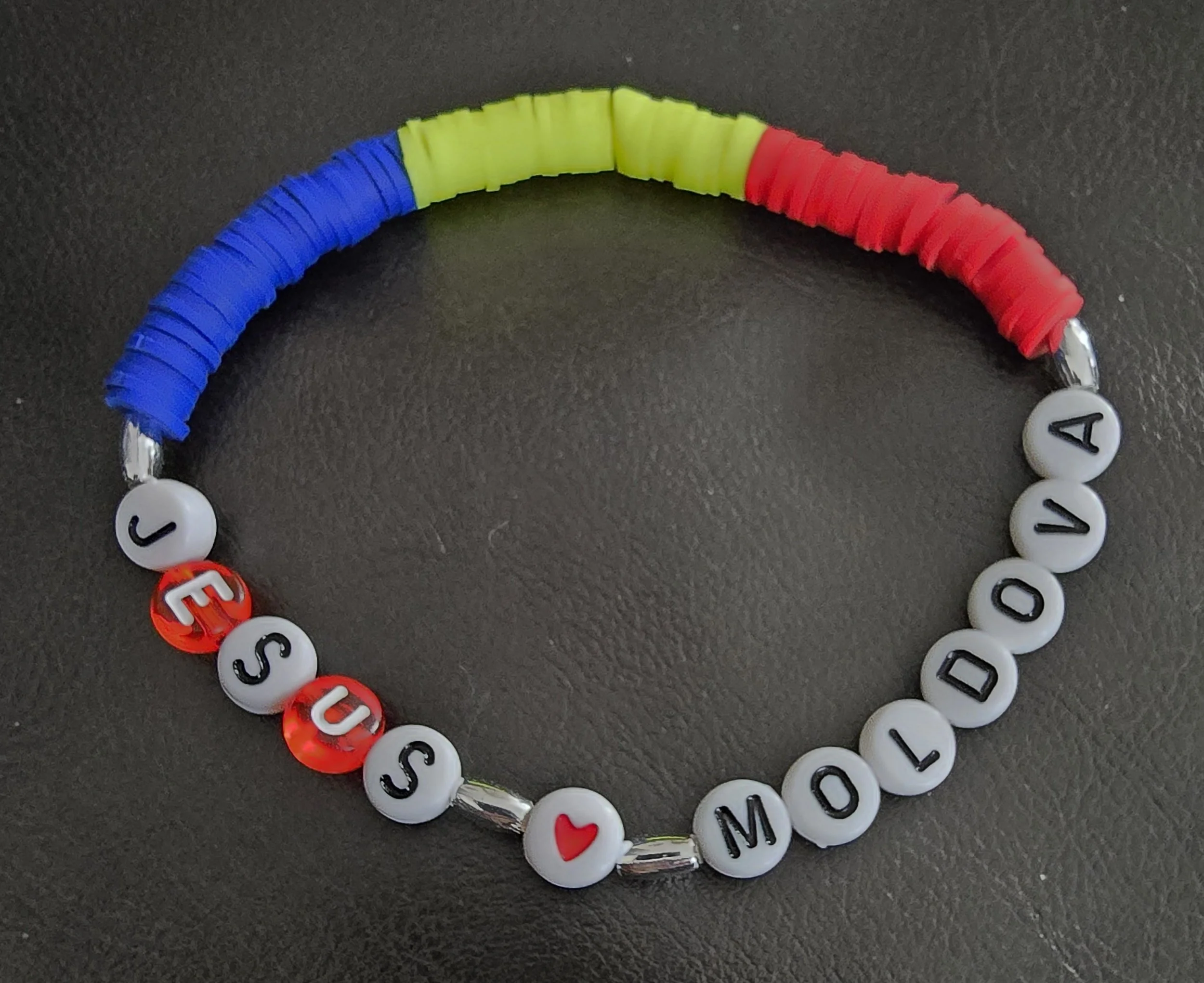 Colorful beaded bracelet spelling out 'JESUS LOVES MOLDOVA' with a heart symbol, on a black surface.