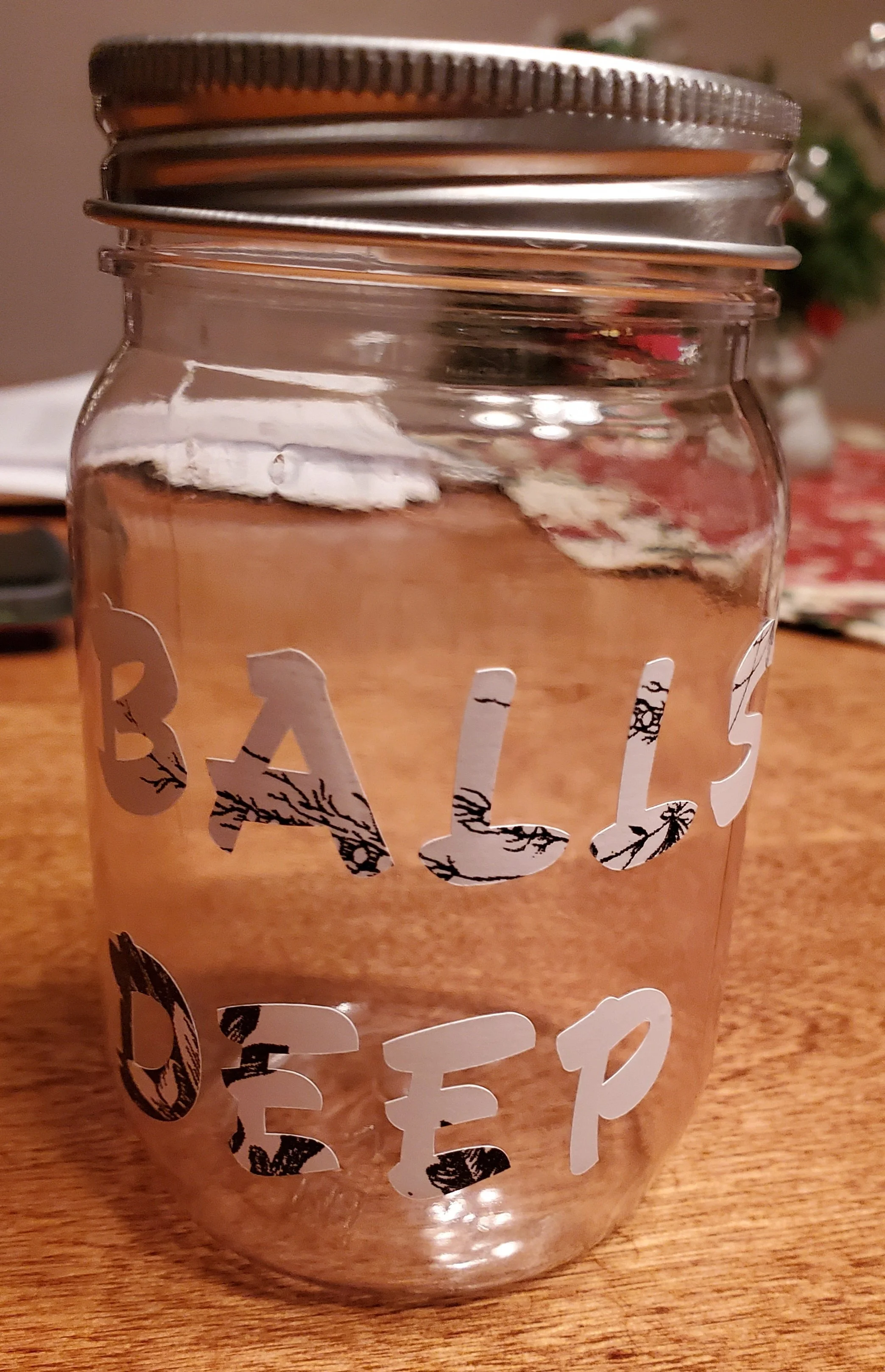 Clear glass jar with a silver lid, decorated with the phrase 'BALLS DEEP' in black and white floral lettering, sitting on a wooden surface.