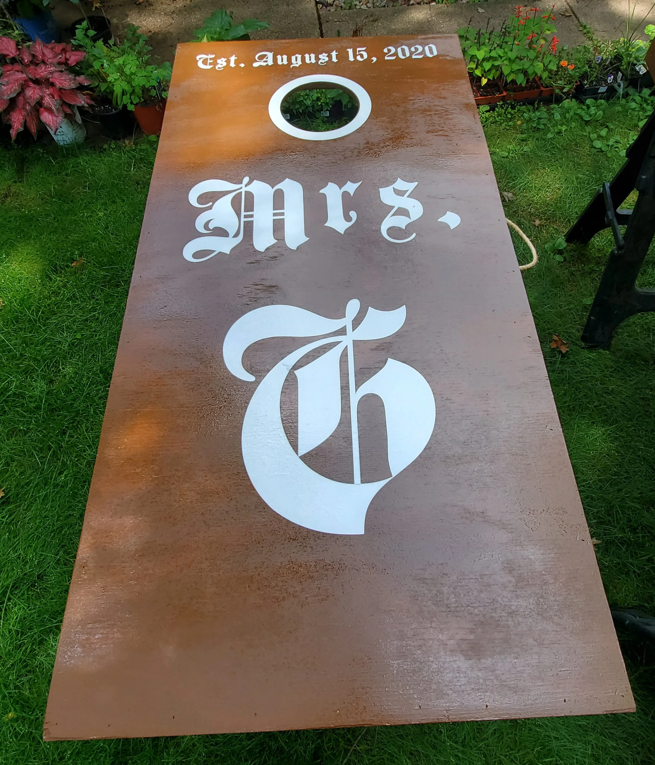 A cornhole board with the word 'Mr.' and the number '6' painted in white on a wooden surface, with a circular hole near the top and grass and potted plants surrounding it.