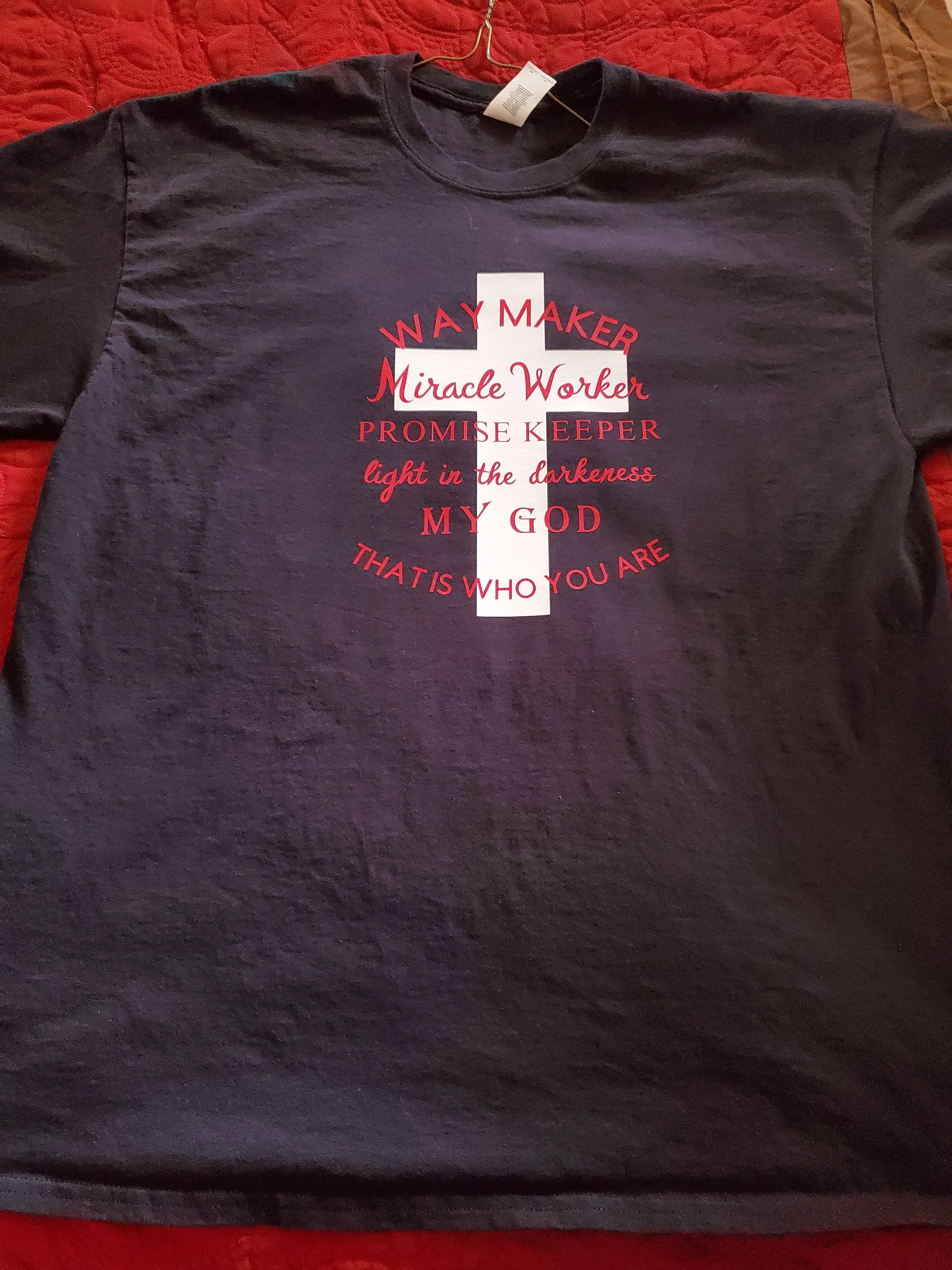 Black T-shirt with a white vertical cross and red text that reads 'Way Maker, Miracle Worker, Promise Keeper, Light in the Darkness, My God, That is Who You Are'.
