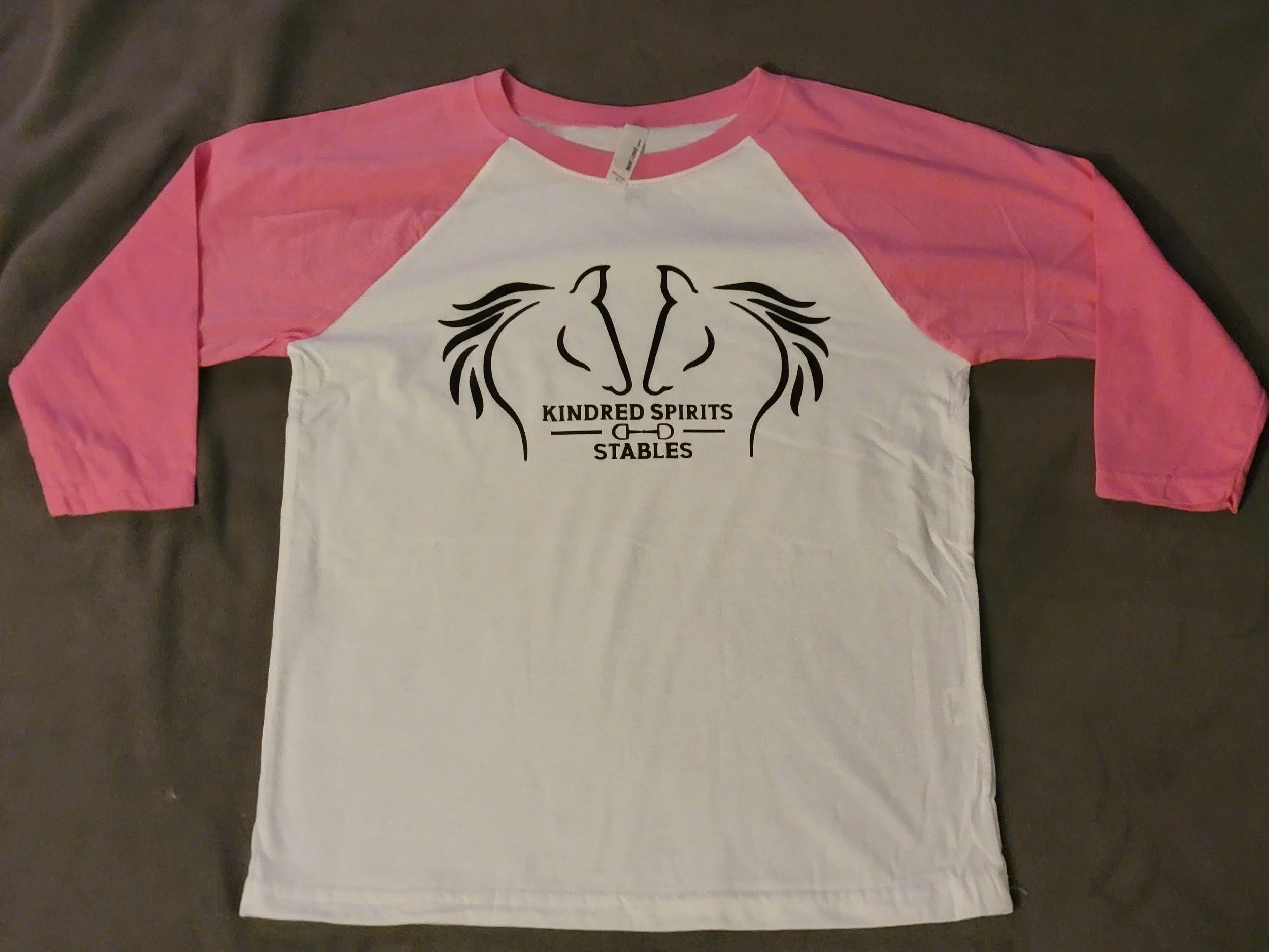White and pink baseball style t-shirt with black logo and text reading "Kindred Spirits & Stables" on the front.