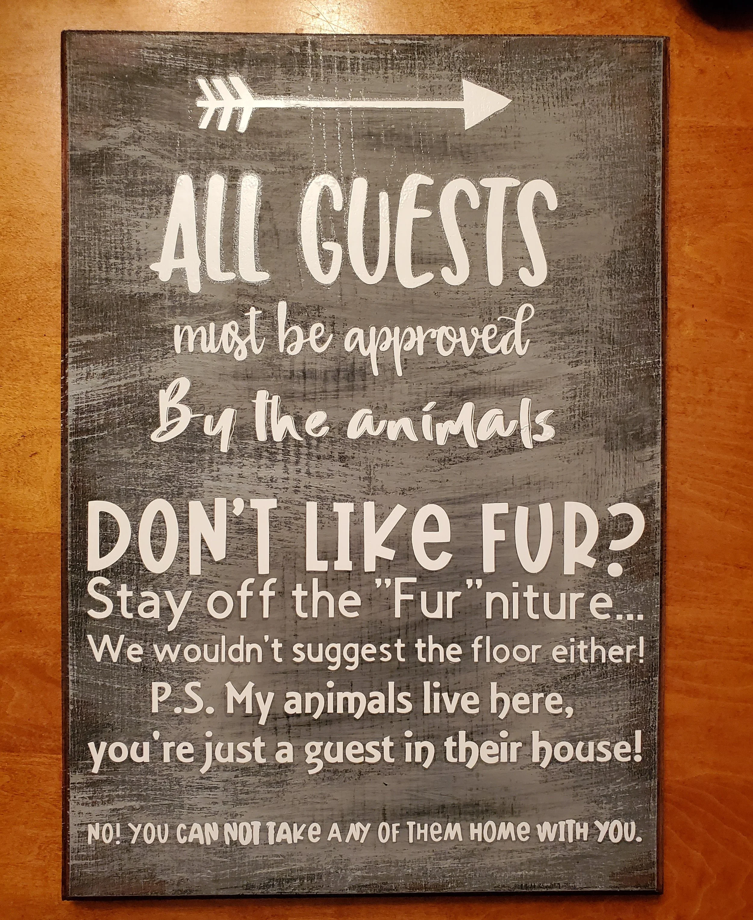 A sign with a rustic wooden background and white text that reads: "All guests must be approved by the animals. Don't like fur? Stay off the 'Fur' niture... We wouldn't suggest the floor either! P.S. My animals live here, you're just a guest in their 